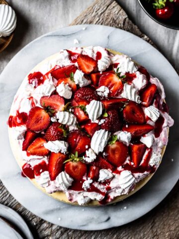 A top view of a vanilla cheesecake topped with whipped cream, strawberry coulis, fresh strawberries and rose water meringues on a marble platter and a rustic wooden cutting board.