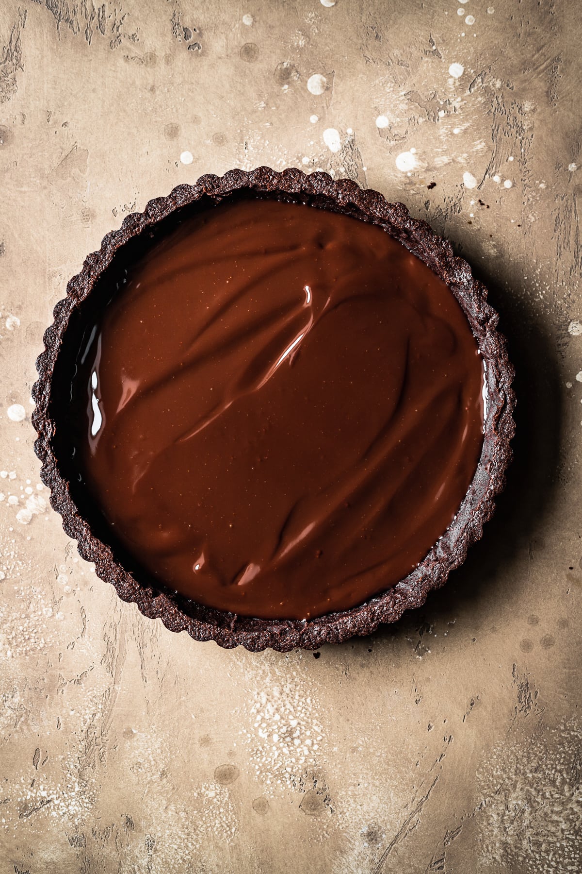 A round chocolate tart shell with a shiny ganache base on a speckled brown backdrop