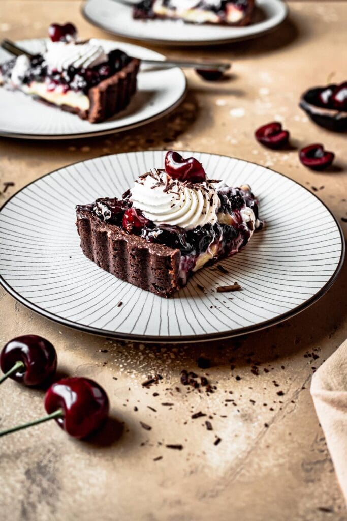 A slice of sour cherry chocolate tart on a plate, focusing on the textures of the tart shell and the piped whipped cream. Another plate is in the background, and cherries are scattered around the brown backdrop.