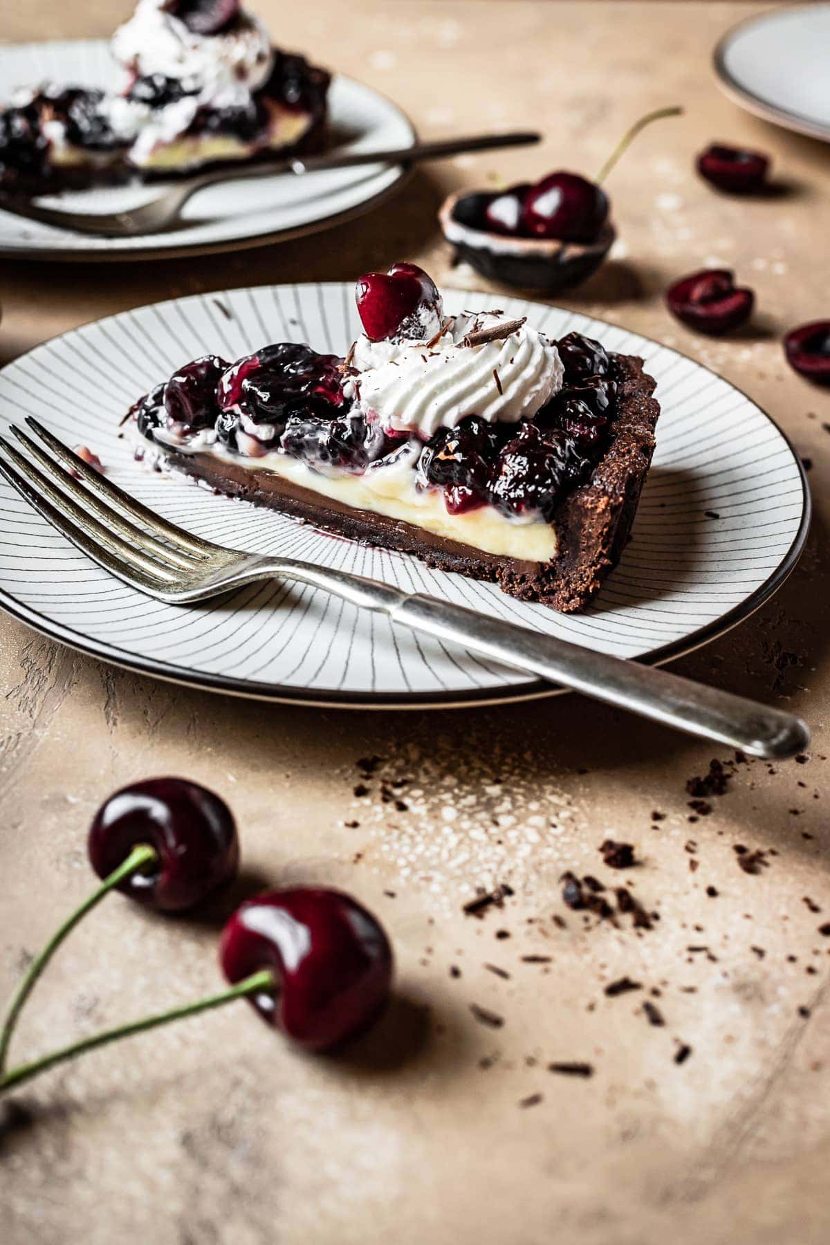 A side view of a slice of tart on a plate. Another plate is in the background, and cherries are scattered around the brown backdrop.