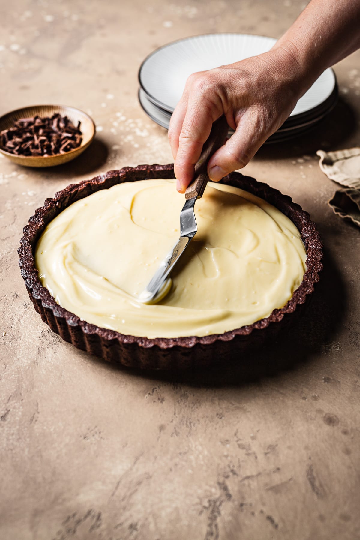 A hand holding a small pastry knife smooths out almond pastry cream into a chocolate tart shell. The scene has a brown background, stack of plates, and a bowl of chocolate shavings nearby.