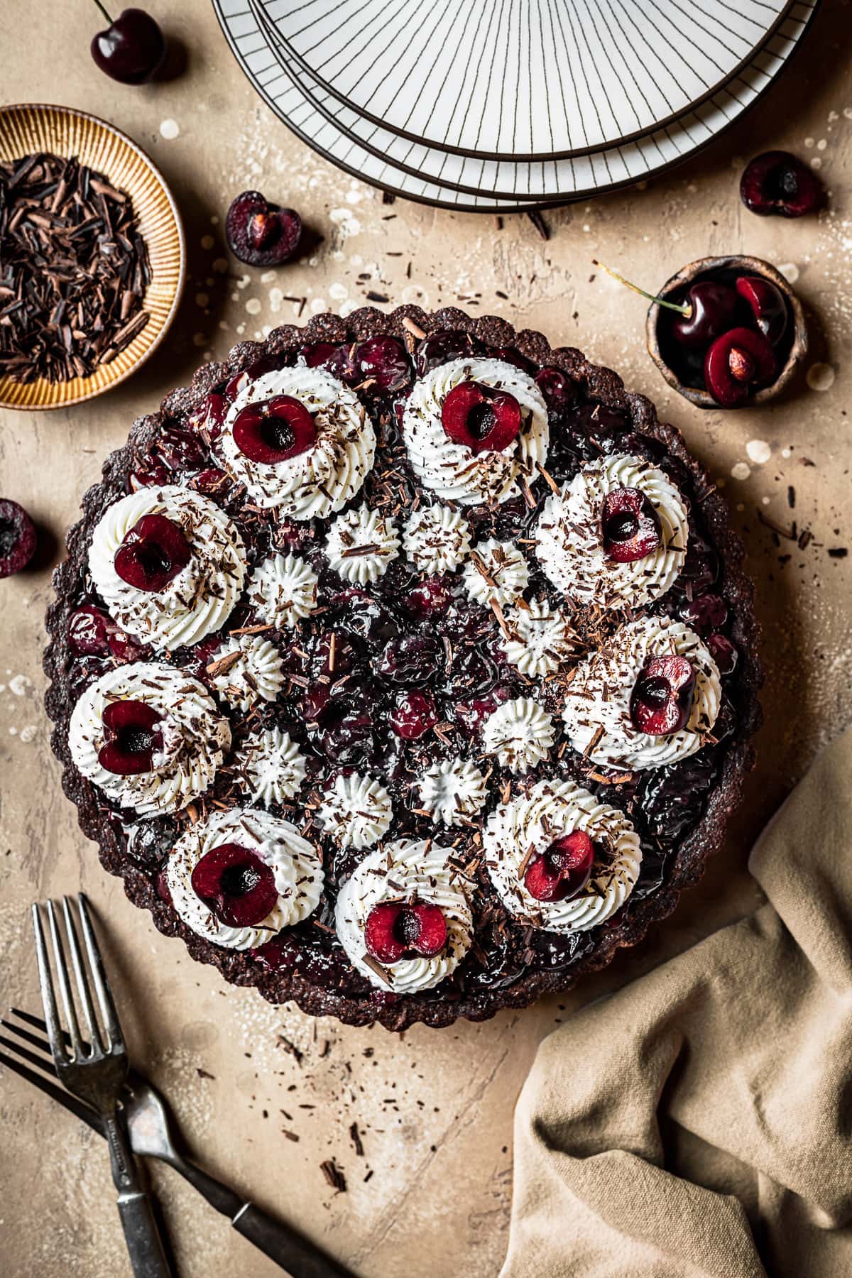 A chocolate cherry tart with piped whipped cream swirls, cherry halves, and chocolate shavings on a brown speckled backdrop. A knife, linen napkin and cherries are placed surrounding the tart.