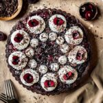 A chocolate cherry tart with piped whipped cream swirls, cherry halves, and chocolate shavings on a brown speckled backdrop. A knife, linen napkin and cherries are placed surrounding the tart.