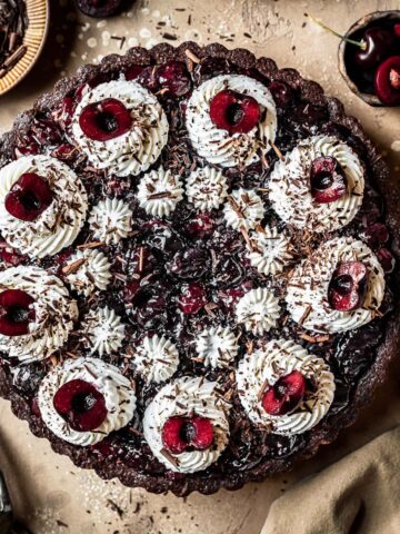 A chocolate cherry tart with piped whipped cream swirls, cherry halves, and chocolate shavings on a brown speckled backdrop. A knife, linen napkin and cherries are placed surrounding the tart.