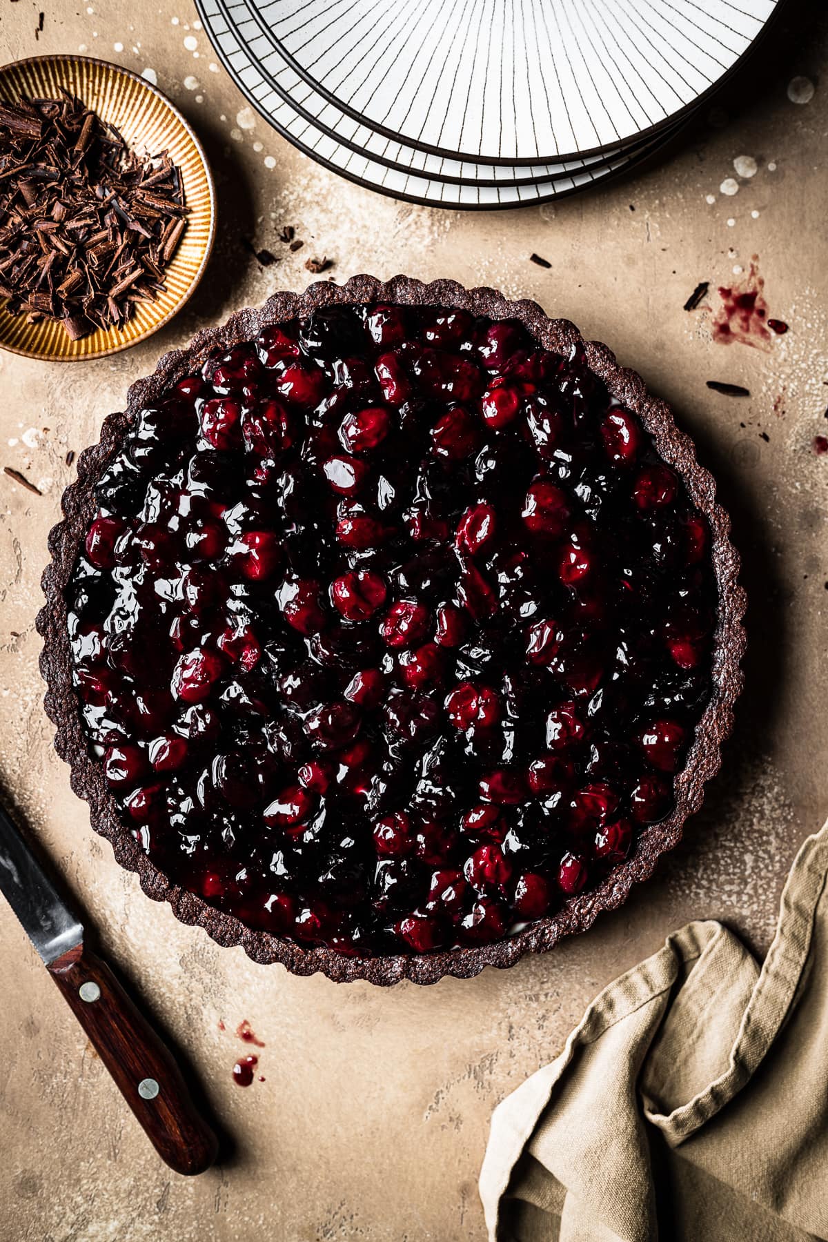 Dark red cherries fill a chocolate crusted tart. The scene has a brown speckled backdrop, a knife, a napkin, stack of plates and a bowl of chocolate shavings nearby.