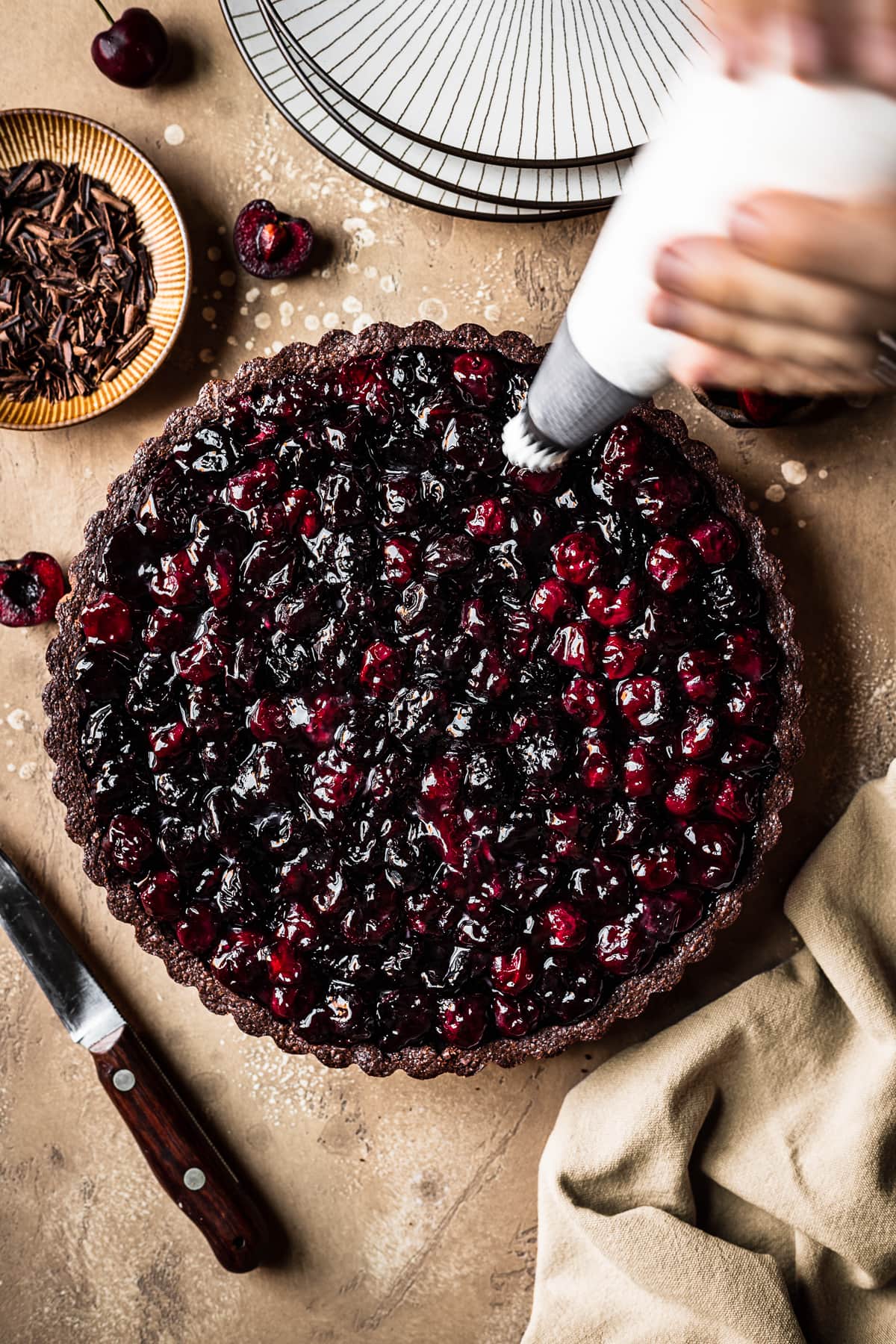 Blurry hands reach into the frame to pipe whipped cream on a chocolate crusted tart. The scene has a brown speckled backdrop, a knife, a napkin, stack of plates and a bowl of chocolate shavings nearby.