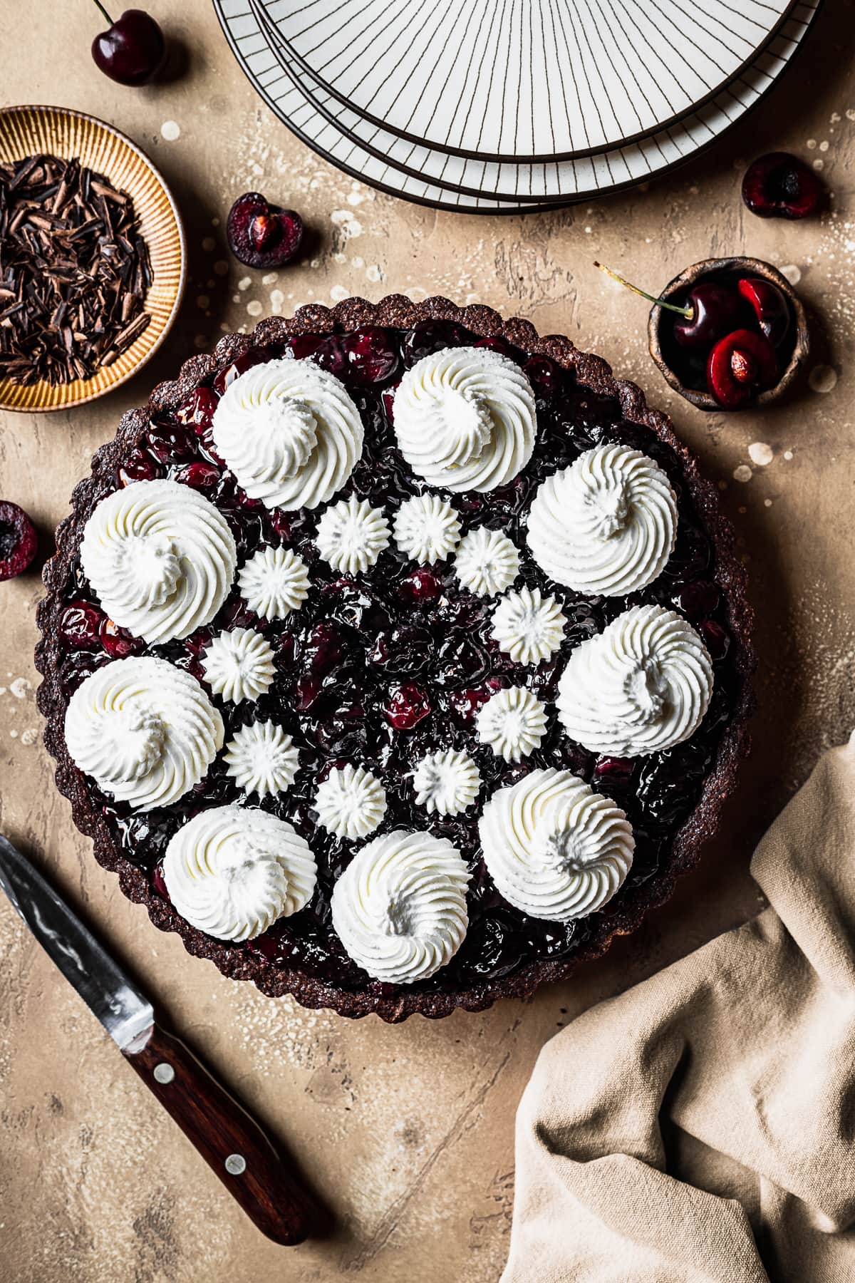 A tart with piped whipped cream swirls on a brown speckled backdrop. A stack of plates, a knife, linen napkin and cherries are placed surrounding the tart.