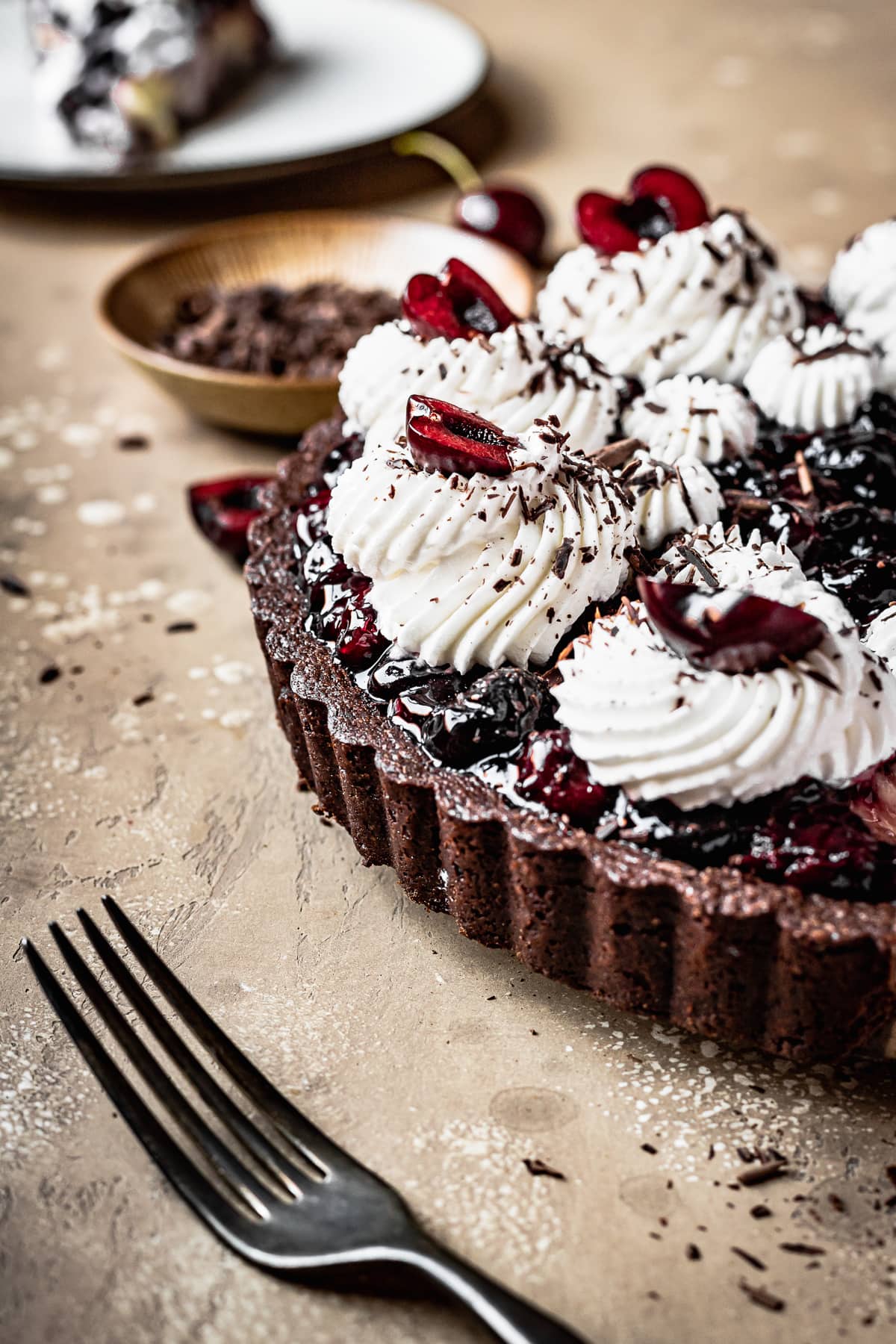 A partial view of a sour cherry tart with chocolate crust on a brown textured backdrop. A bowl of chocolate shavings is in the background along with an additional slice of tart.