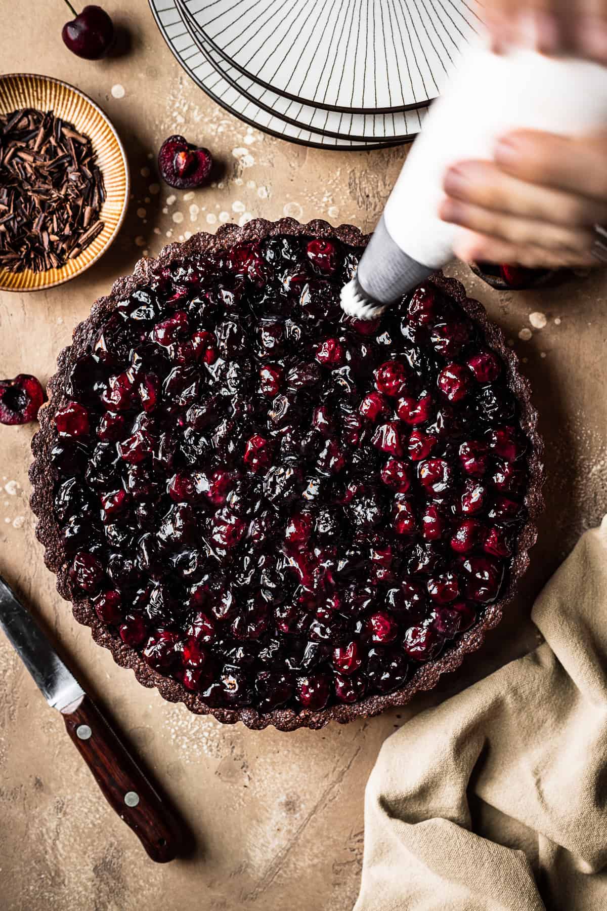 A cherry chocolate tart with hands holding a piping bag filled with whipped cream.