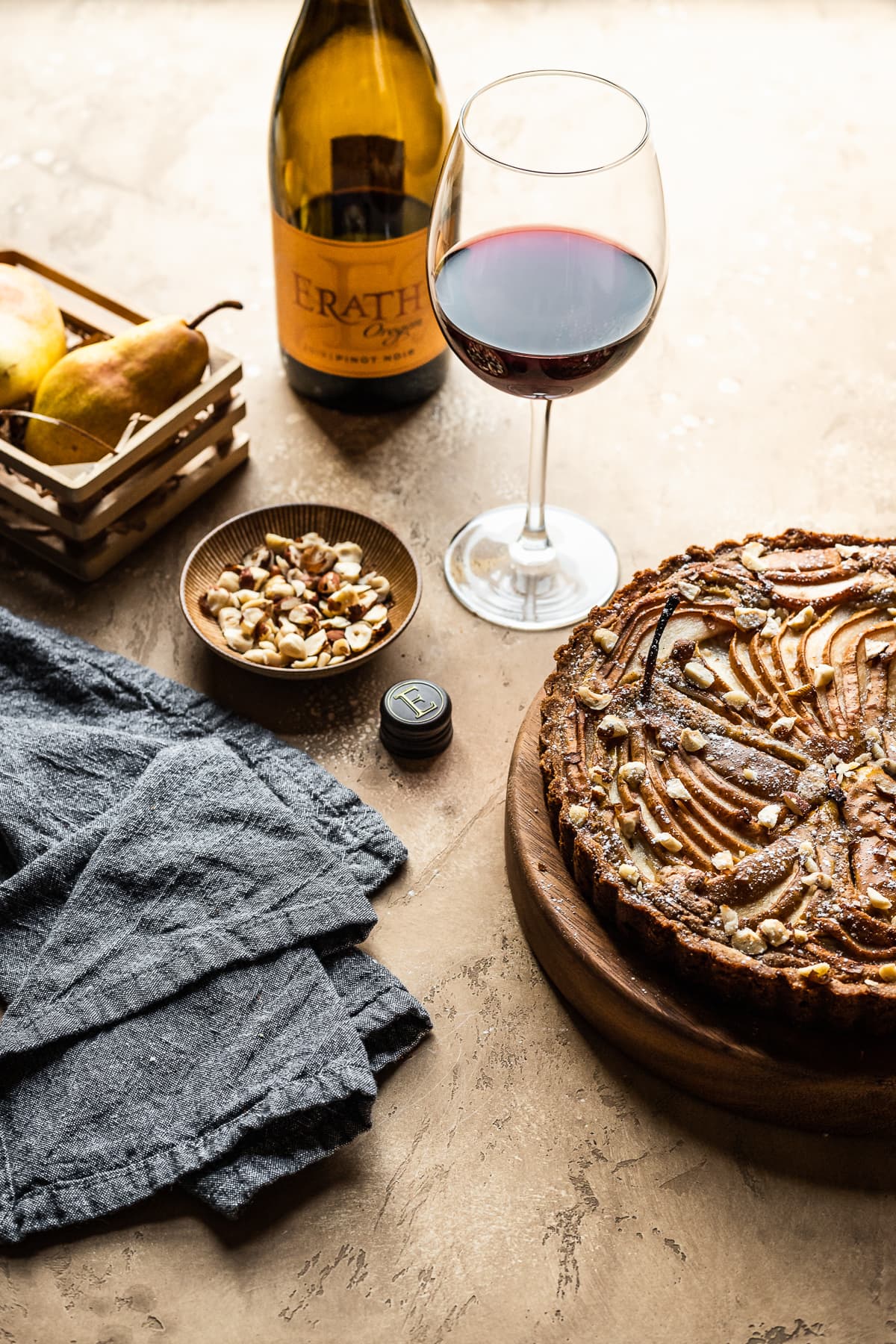 A pear tart on a wooden platter on a warm tan textured background. Nearby is a small bowl of nuts, a grey textured napkin, a small wooden crate of pears, and a glass of red wine with bottle.