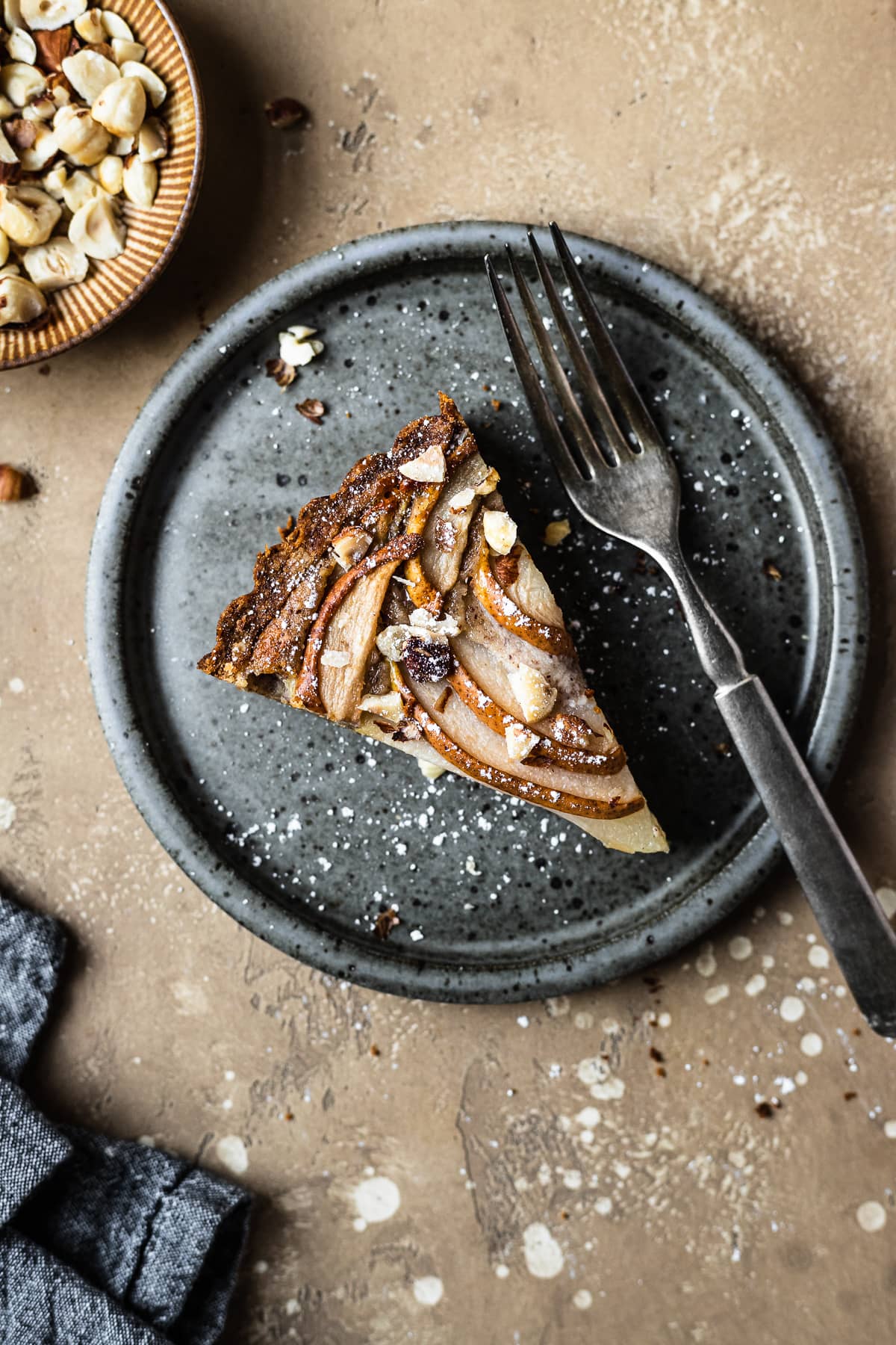 Pear hazelnut tart slice on a speckled grey blue ceramic plate. A vintage fork rests on the right side of the plate. The plate is on a warm tan textured stone background. A small bit of a textured grey napkin peeks out from the bottom left corner, and a small view of a bowl of chopped hazelnuts is visible at top left.