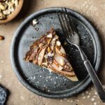 Pear hazelnut tart slice on a speckled grey blue ceramic plate. A vintage fork rests on the right side of the plate. The plate is on a warm tan textured stone background. A small bit of a textured grey napkin peeks out from the bottom left corner, and a small view of a bowl of chopped hazelnuts is visible at top left.