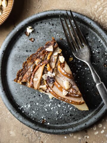 Pear hazelnut tart slice on a speckled grey blue ceramic plate. A vintage fork rests on the right side of the plate. The plate is on a warm tan textured stone background. A small bit of a textured grey napkin peeks out from the bottom left corner, and a small view of a bowl of chopped hazelnuts is visible at top left.