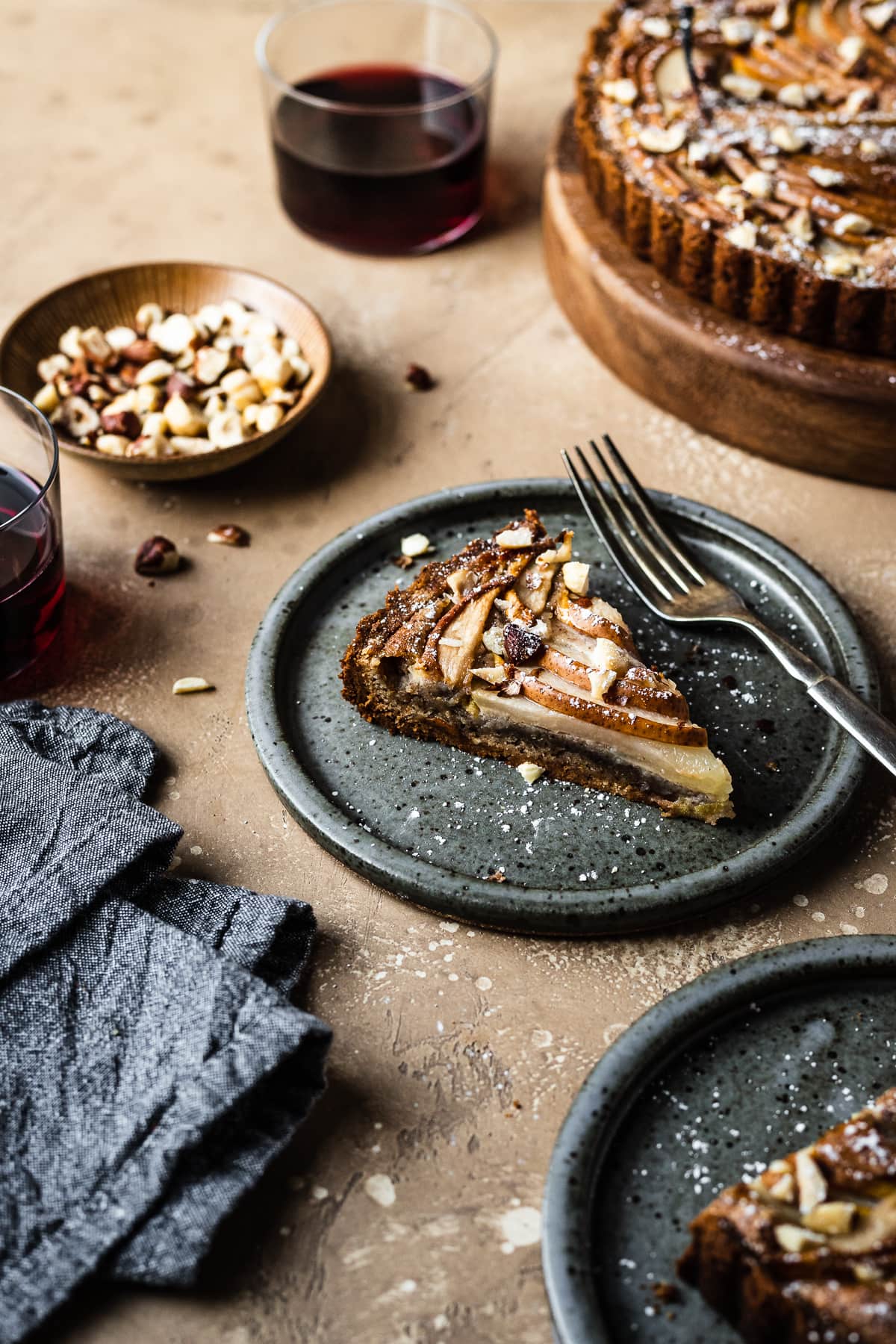 An angled view of two slices of tart on speckled grey blue ceramic plates. Nearby is the remainder of the tart, a small bowl of hazelnuts, and two stemless wine glasses filled with red wine. A grey textured linen napkin is at bottom left. The background of the image is a warm tan speckled stone.