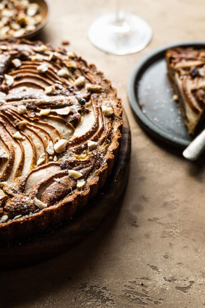 A baked pear hazelnut tart peeks into view from the left side of the image, shot at a 45 degree angle. Golden light is shining on the tart from behind. A slice of tart sits on a ceramic plate in the background, along with a small bowl of chopped nuts and a partial view of a wine glass stem.