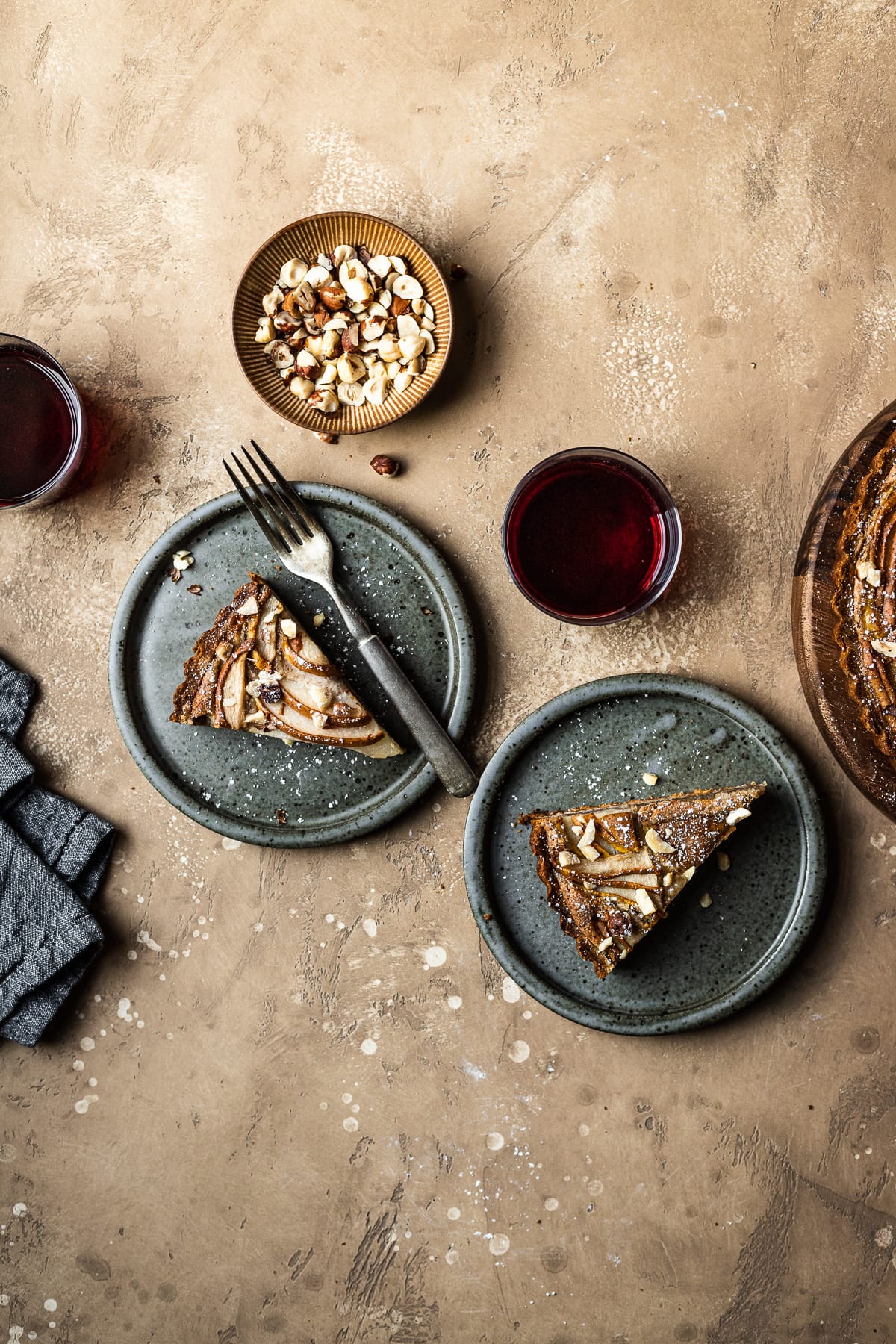 Two slices of tart on speckled grey blue ceramic plates. To the right is the remainder of the tart. A small bowl of hazelnuts, and two stemless wine glasses filled with red wine surround the plates. A grey textured linen napkin is at lower left. The background of the image is a warm tan speckled stone.