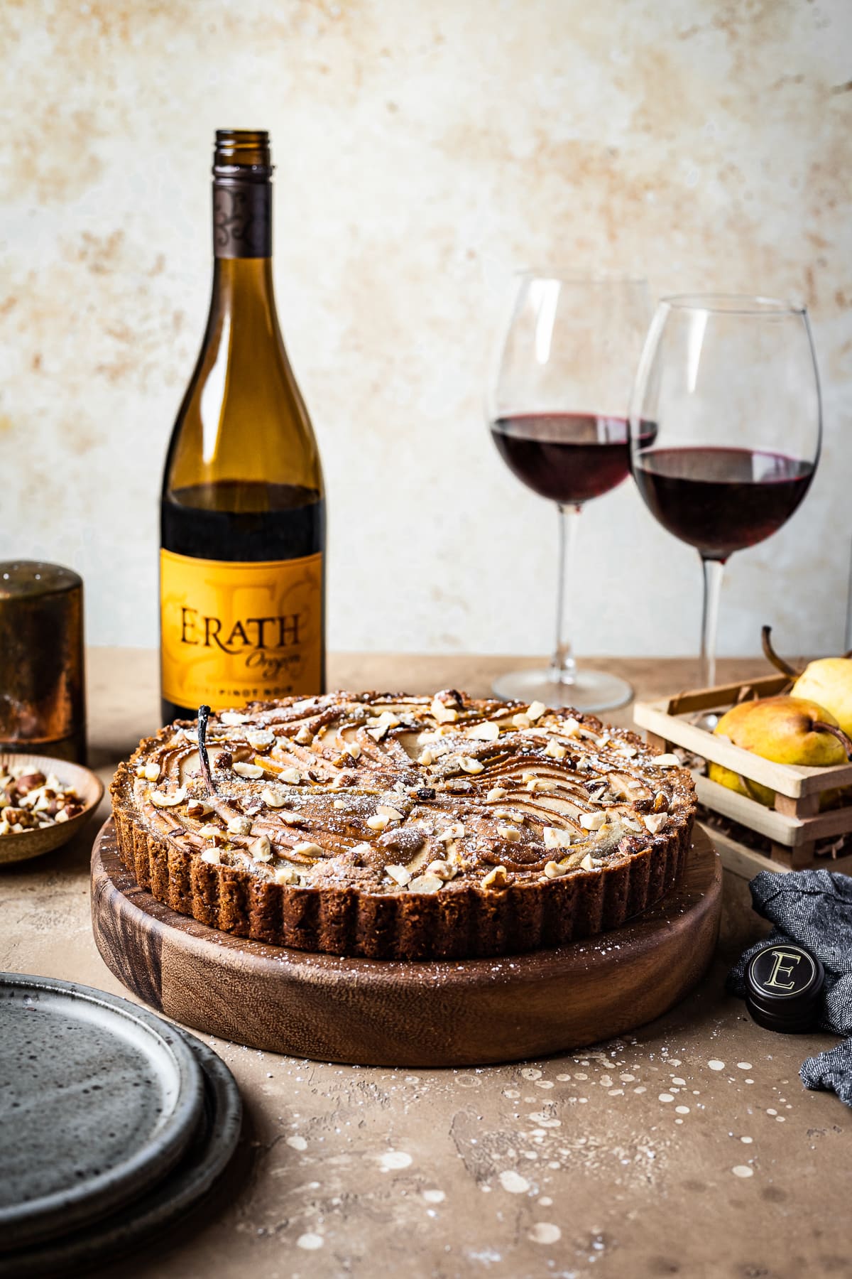 A front view of a pear tart on a round wooden platter on a textured warm tan background. The tart is surrounded by blue grey ceramic plates, an opened bottle of red wine, two half full glasses of red wine, a small wooden crate of pears, and a textured grey napkin.