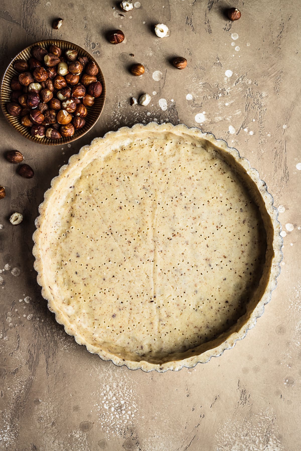 An unbaked round tart crust with a small bowl of hazelnuts at top left, resting on a textured speckled tan background.