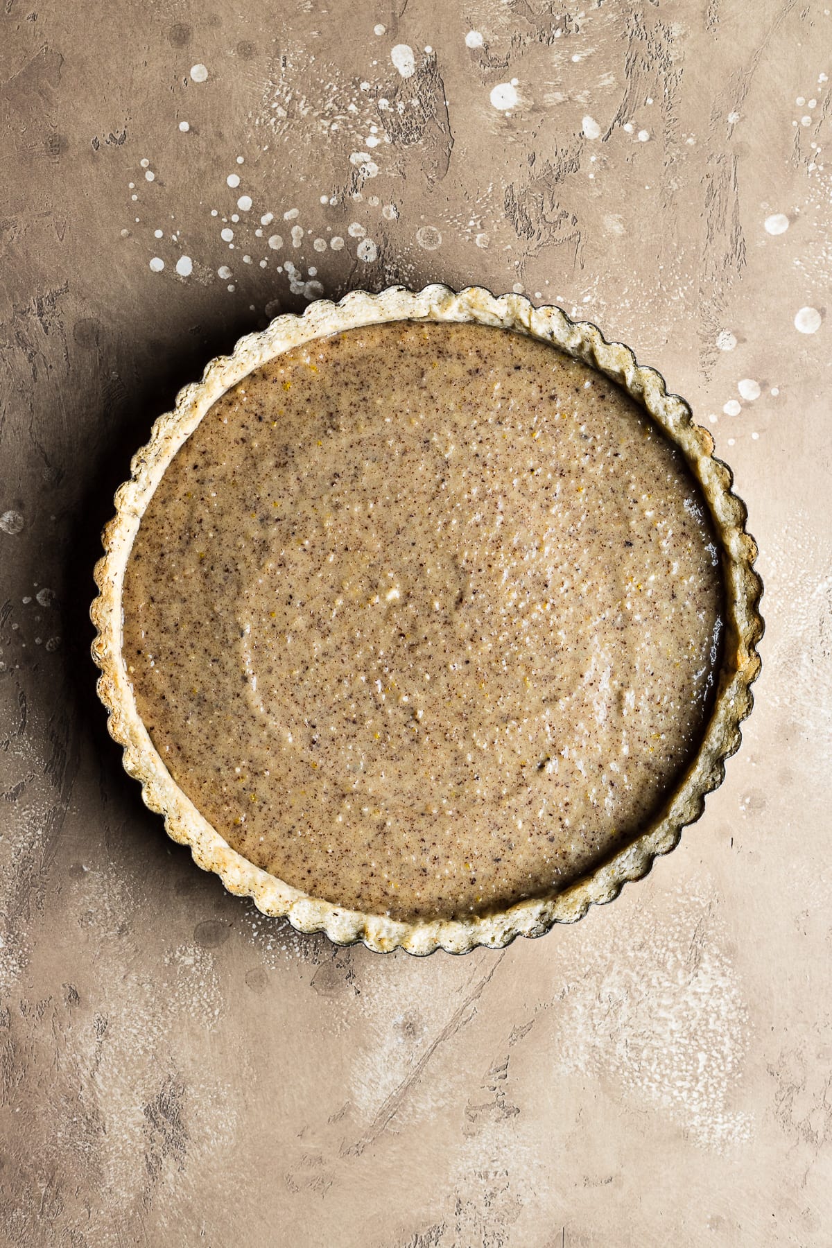 A blind baked round tart crust filled with hazelnut frangipane resting on a textured speckled tan background.