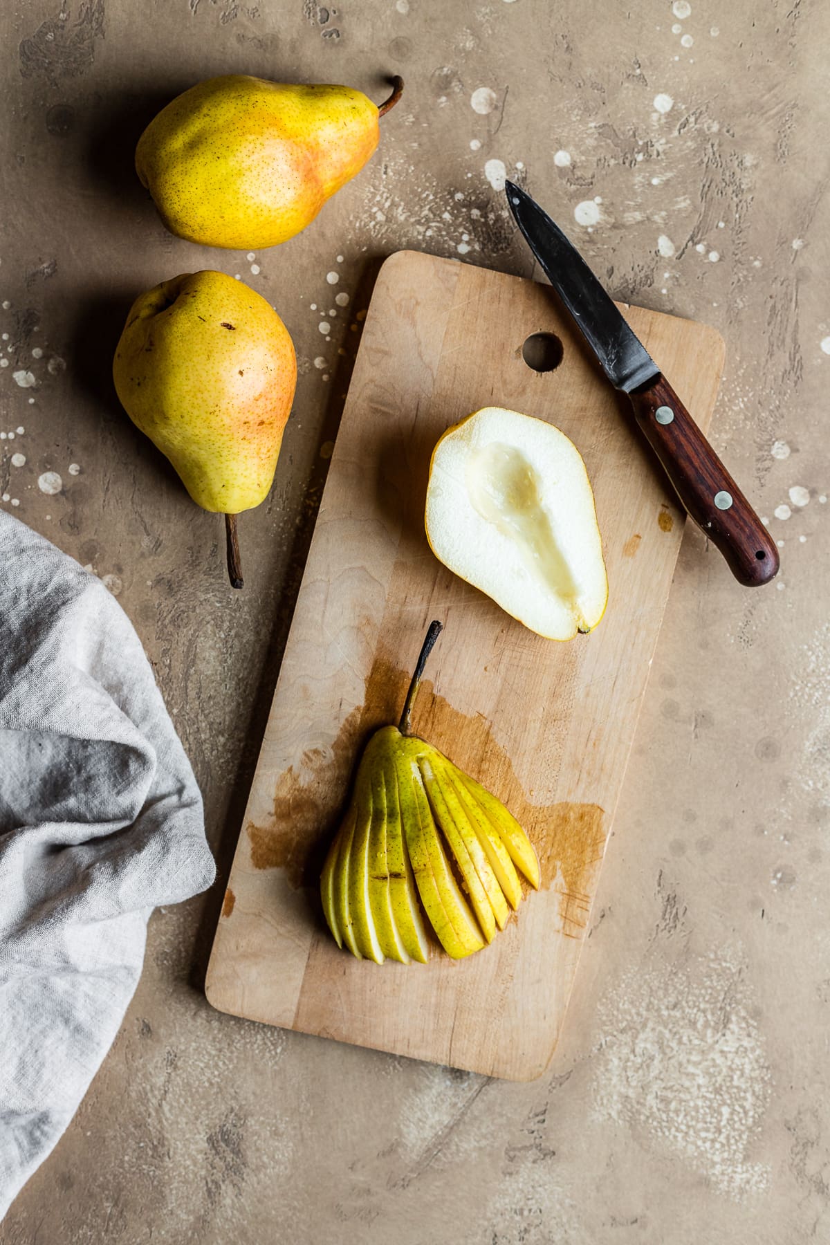 A wooden cutting board with two pear halves resting on it. One is face up with the core removed, and another is face down and has been cut into a fan shape with thin slices but the top of the pear intact. A wooden knife rests on the board, and two whole pears are at top left. A pale linen napkin is at bottom left. The scene has a textured tan stone background.