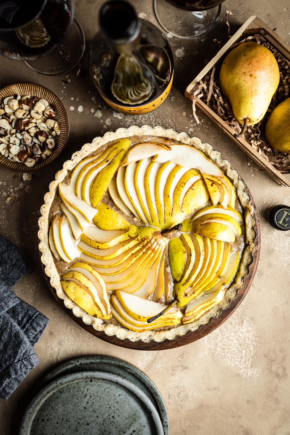 An unbaked pear hazelnut tart in a round pan is at the center of the image. Surrounding the tart are speckled blue ceramic plates, a bowl of chopped hazelnuts, pears in a wooden crate, a linen napkin, and a bottle of red wine with two filled wine glasses. Everything rests on a textured tan background.