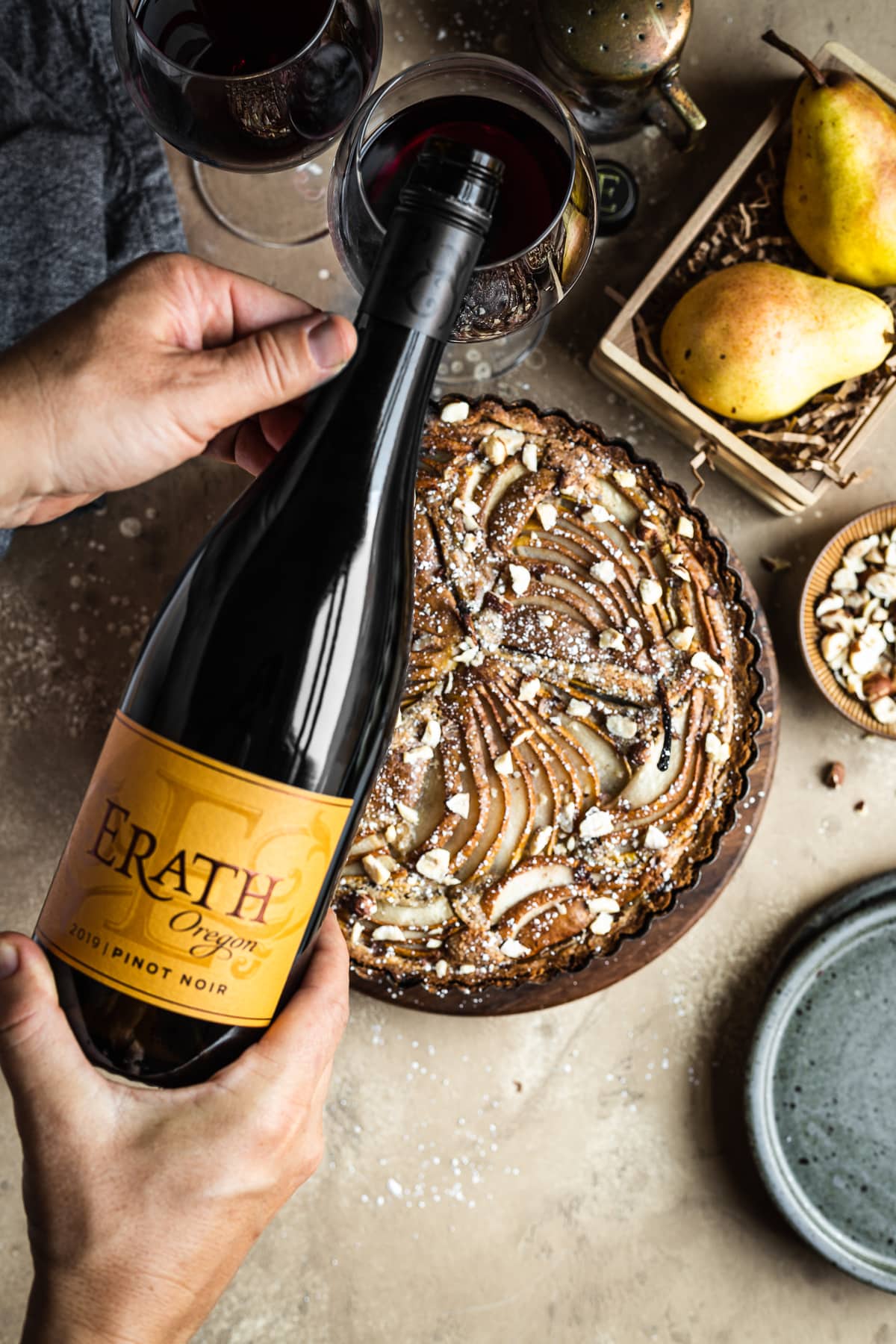 Hands pouring a bottle of red wine into glasses with a golden brown pear tart in the background. Surrounding the tart are speckled blue ceramic plates, a bowl of chopped hazelnuts, pears in a wooden crate, and a linen napkin. Everything rests on a textured tan background.