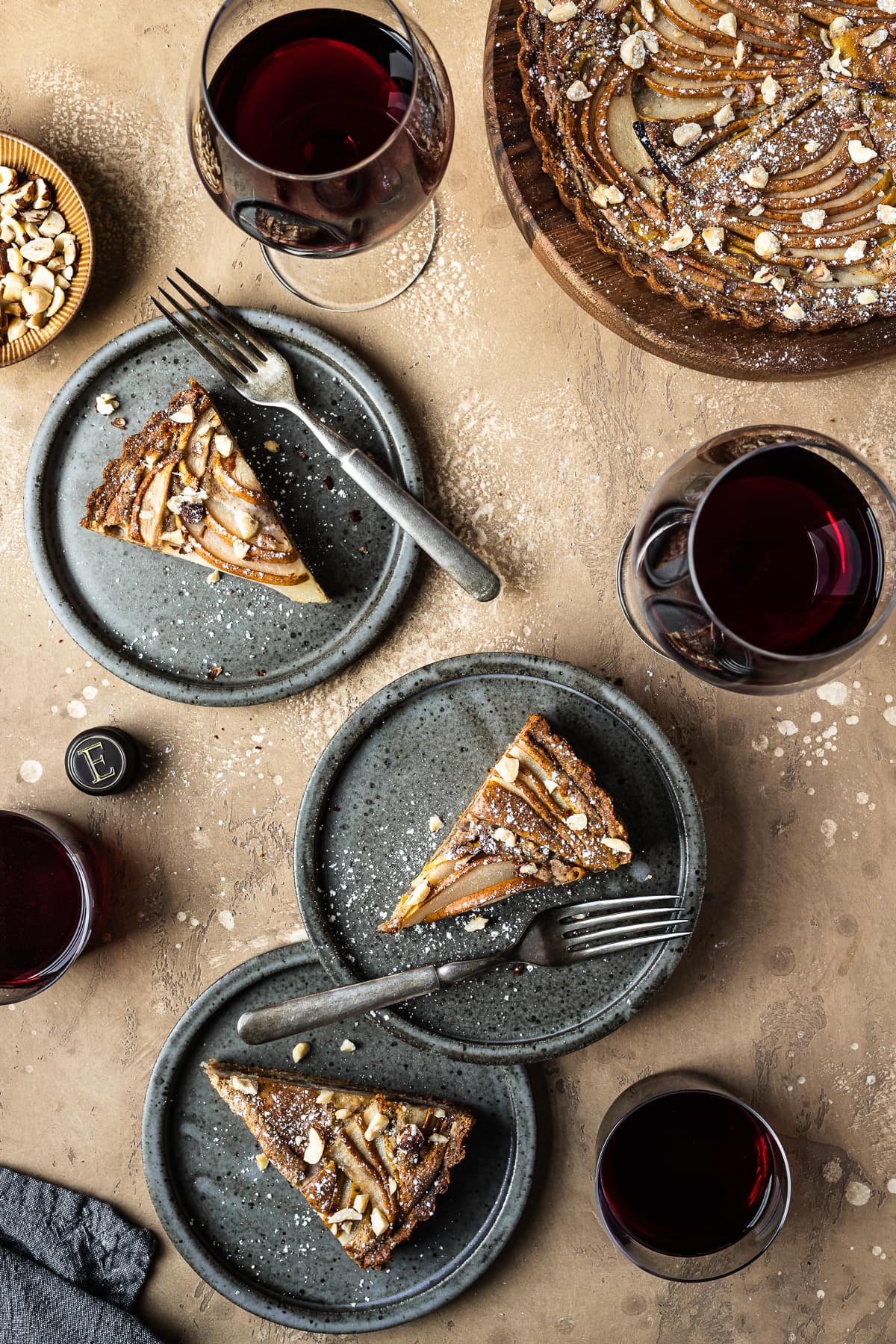 Three pear hazelnut tart slices on speckled grey blue ceramic plates. Vintage forks rests on the right side of the slices. The plates are on a warm tan textured stone background. A small bit of a textured grey napkin peeks out from the bottom left corner, and a small view of a bowl of chopped hazelnuts is visible at top left. There are four full glasses of red wine arranged throughout the image. The remainder of the tart is at top right.