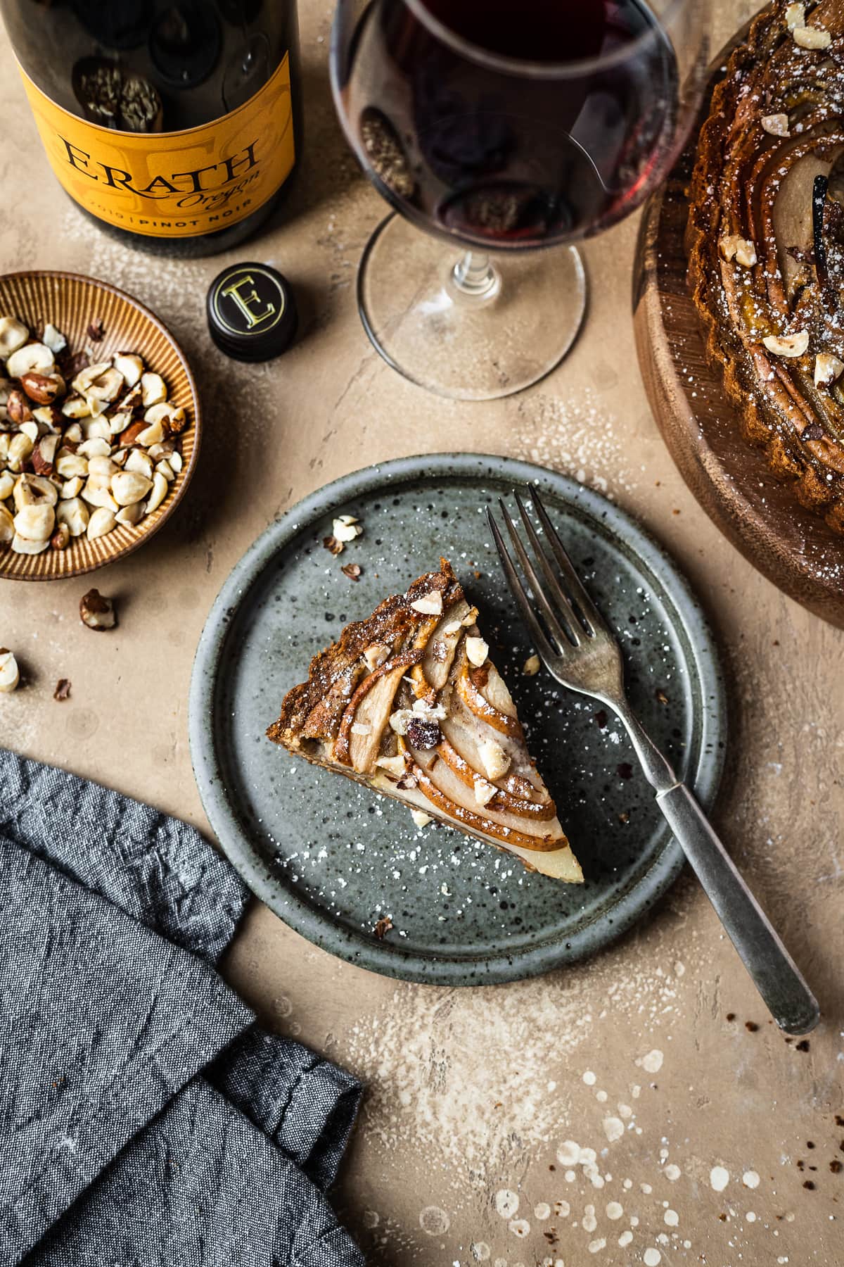 Pear hazelnut tart slice on a speckled grey blue ceramic plate. A vintage fork rests on the right side of the plate. The plate is on a warm tan textured stone background. A textured grey napkin is at the bottom left corner, and a small bowl of chopped hazelnuts is visible at top left along with a wine bottle and a glass of red wine at top. The remainder of the tart peeks out at top right.