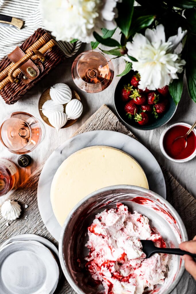 A picnic scene showing a vanilla cheesecake with hands holding a bowl of strawberry swirled whipped cream above it. Surrounding the cheesecake are wine bottles, a picnic basket, meringues, a bowl of strawberries and a bouquet of flowers. 