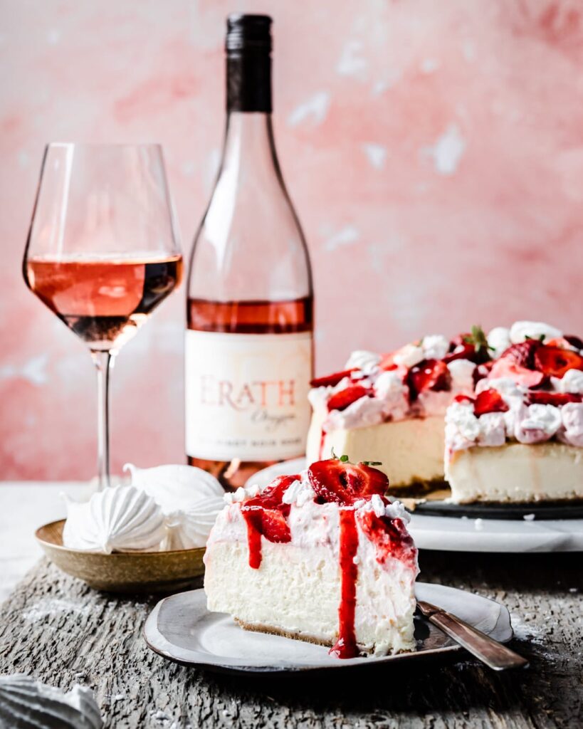A slice of vanilla cheesecake topped with whipped cream and strawberries. In the background there is a bottle of rose wine and a wine glass half full of rose. The platter of cheesecake rests in the background on a marble platter. The background is a light coral pink.