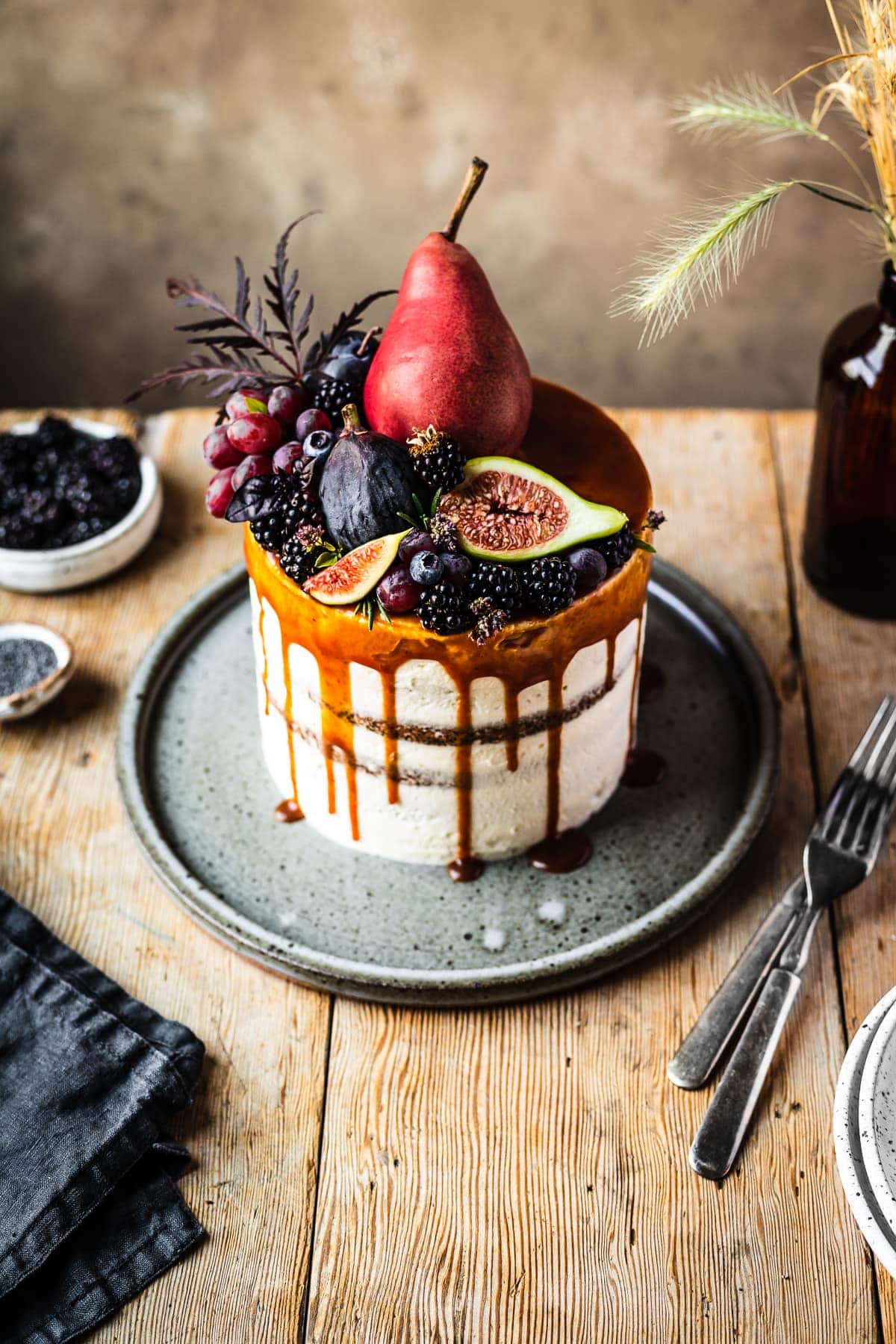 A 45 degree angle view of a layer cake with a caramel drip and a crown of late summer fruit on top. The cake sits on a blue grey ceramic platter on a rustic wooden table with forks, a dark blue linen napkin, and small bowls of poppy seeds and blackberries nearby. There is a warm tan stone background behind the table.