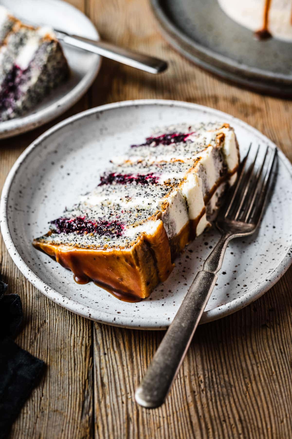A slice of poppyseed cake with mascarpone frosting and caramel drip, with layers of vibrant purple blackberry basil jam. The slice sits on a white speckled ceramic plate on a rustic wooden table.