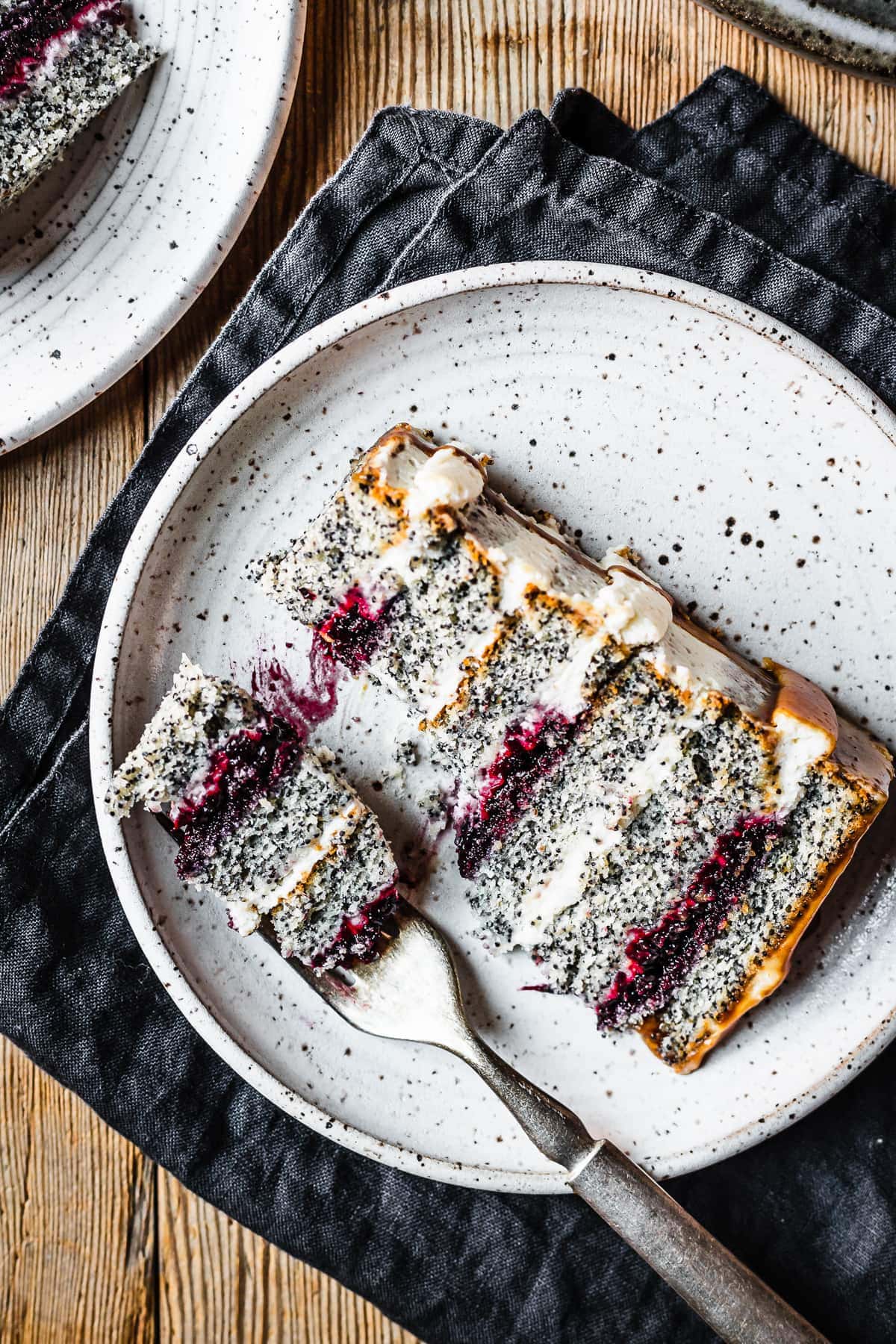 A slice of poppyseed cake with mascarpone frosting and caramel drip, with layers of vibrant purple blackberry basil jam. The slice sits on a white speckled ceramic plate with a navy blue linen napkin folded underneath, on a rustic wooden table.