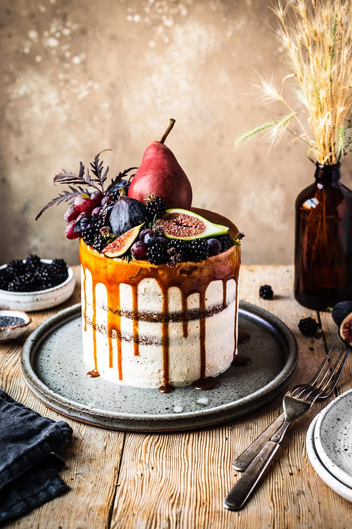 A white frosted cake with a caramel drip and a crown of late summer fruit on top. The cake sits on a blue grey ceramic platter on a rustic wooden table with forks and small bowls of poppy seeds and blackberries nearby. There is a warm tan stone background behind the table.