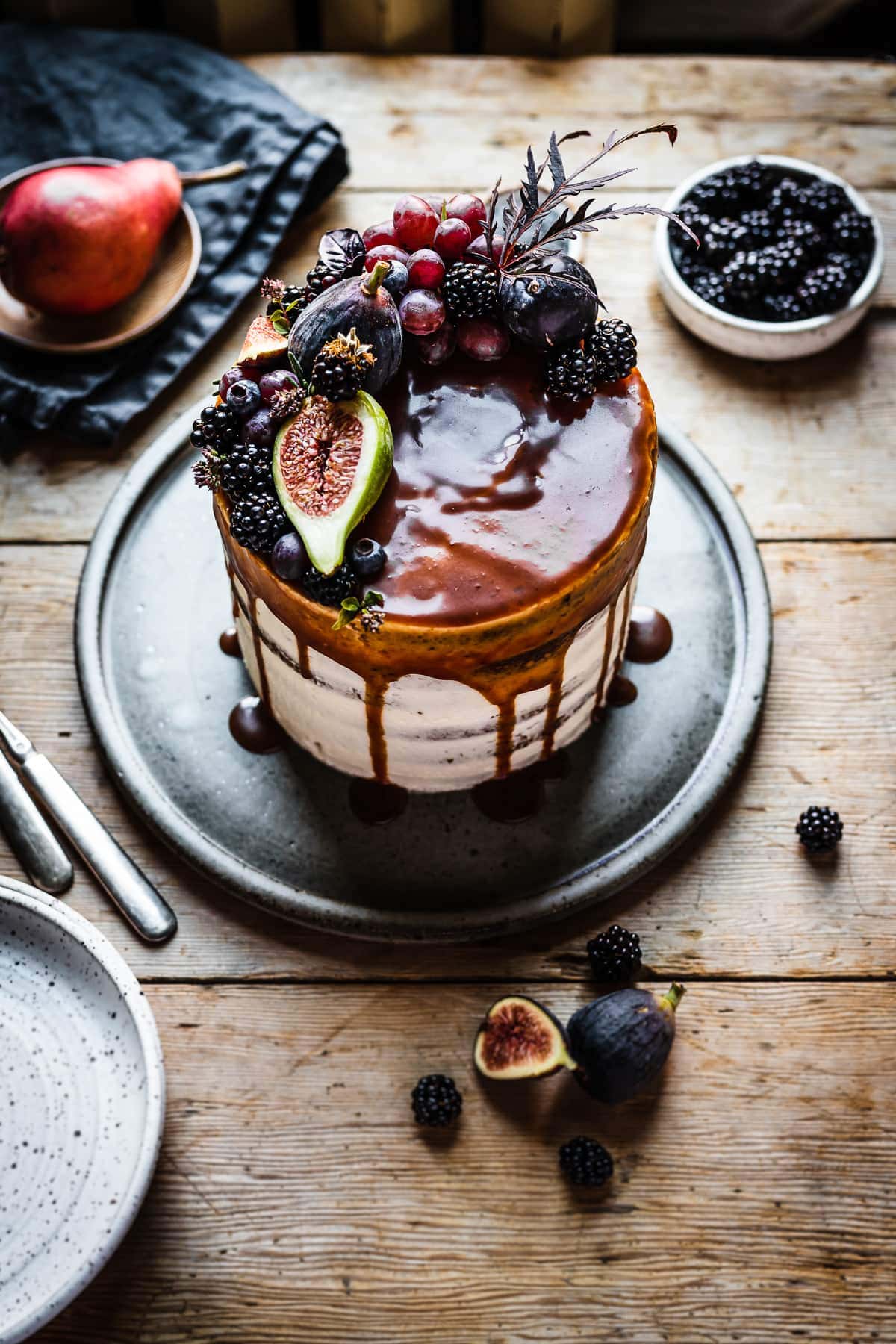 A white frosted layer cake with a caramel drip and a crown of late summer fruit on top. The cake sits on a blue grey ceramic platter on a rustic wooden table with forks and small bowls of poppy seeds and blackberries nearby. The light is coming from behind the cake and makes the caramel on top of the cake shine.