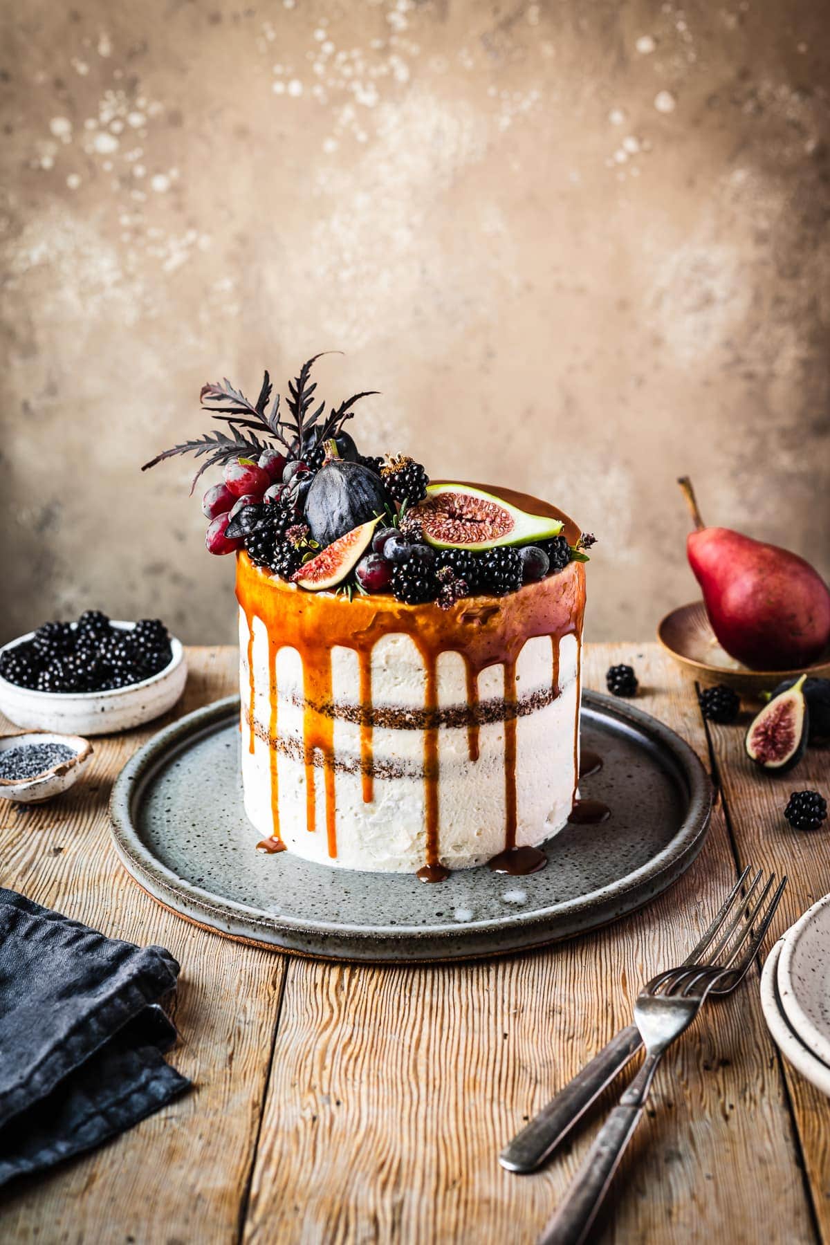 A white frosted blackberry poppy seed layer cake with a caramel drip and a crown of late summer fruit on top. The cake sits on a blue grey ceramic platter on a rustic wooden table with forks and small bowls of poppy seeds and blackberries nearby. There is a warm tan stone background behind the table.