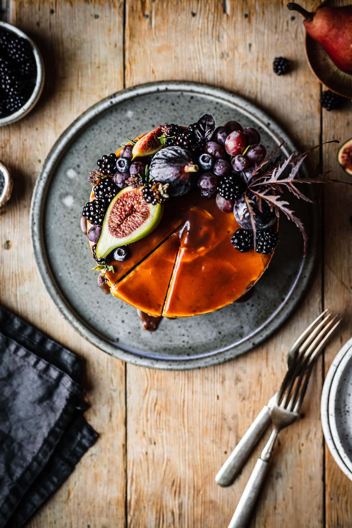 Top view of a cake topped with caramel and a crescent shaped crown of late summer fruit, including figs, blackberries and grapes. A slice of cake has been cut but not removed. The cake sits on a blue grey ceramic platter on a rustic wooden table with forks, a dark blue napkin, and small bowls of poppy seeds and blackberries nearby.