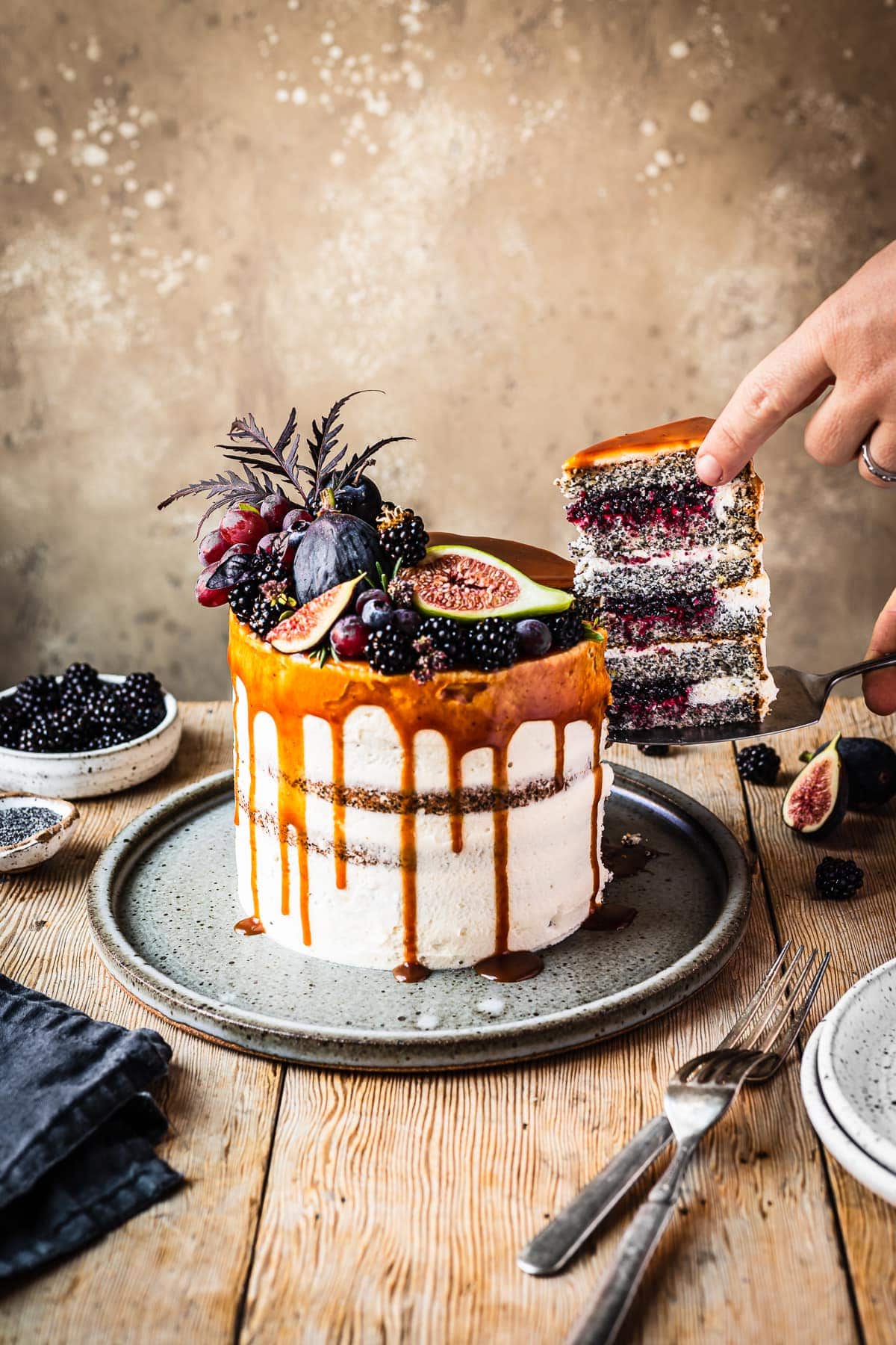 A white frosted layer cake with a caramel drip and a crown of late summer fruit on top. Hands are removing a slice balanced on a cake server, revealing layers of poppy seed cake, vibrant purple jam and white frosting. The cake sits on a blue grey ceramic platter on a wooden table with forks and small bowls of poppy seeds and blackberries nearby. There is a warm tan stone background behind the table.