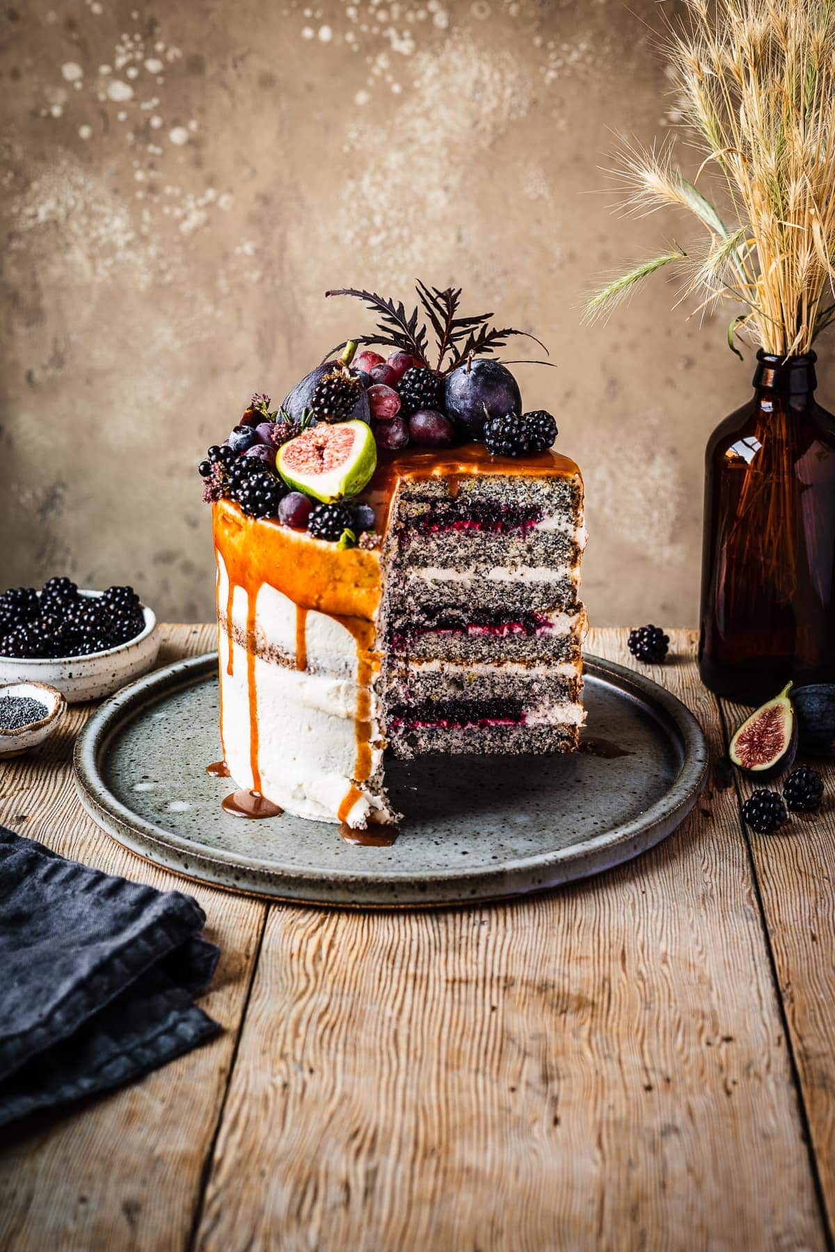 A cross section of a white layer cake with a caramel drip and a crown of late summer fruit on top. Slices are missing, revealing layers of poppy seed cake, vibrant purple jam and white frosting. The cake sits on a blue grey ceramic platter on a wooden table with forks and small bowls of poppy seeds and blackberries nearby. There is a warm tan stone background behind the cake and table.