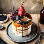 A white layer cake with a caramel drip and a crown of late summer fruit on top. The cake sits on a blue grey ceramic platter on a wooden table with forks and small bowls of poppy seeds and blackberries nearby.