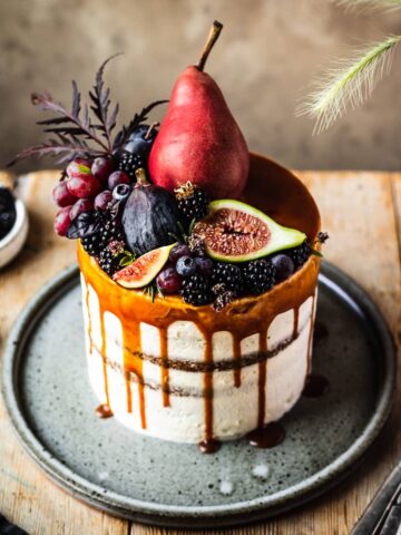 A white layer cake with a caramel drip and a crown of late summer fruit on top. The cake sits on a blue grey ceramic platter on a wooden table with forks and small bowls of poppy seeds and blackberries nearby.