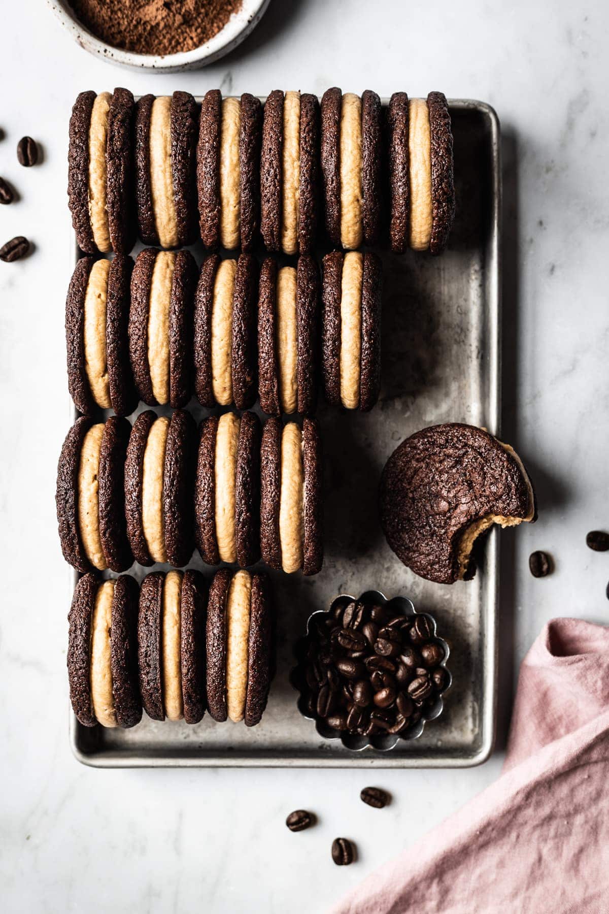 A silver metal cookie sheet holds 4 tidy rows of frosting filled sandwich cookies on end to show their centers. One is lying flat with a bite taken out of it. The cookie sheet is on a white marble surface. In one corner of the cookie sheet there is a small fluted container of coffee beans and a pink linen napkin at bottom right. At top left there is a portion of a small white ceramic bowl filled with cocoa powder visible.