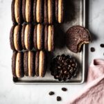 A silver metal cookie sheet holds 3 tidy rows of frosting filled sandwich cookies on end to show their centers. One is lying flat with a bite taken out of it. The cookie sheet is on a white marble surface. In one corner of the cookie sheet there is a small fluted container of coffee beans and a pink linen napkin at bottom right.