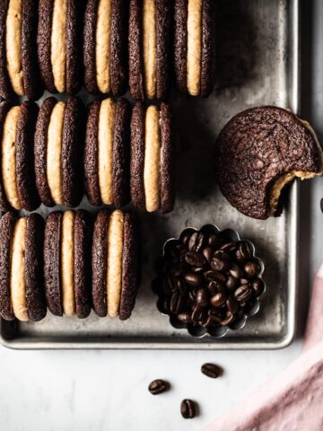 A silver metal cookie sheet holds 3 tidy rows of frosting filled sandwich cookies on end to show their centers. One is lying flat with a bite taken out of it. The cookie sheet is on a white marble surface. In one corner of the cookie sheet there is a small fluted container of coffee beans and a pink linen napkin at bottom right.