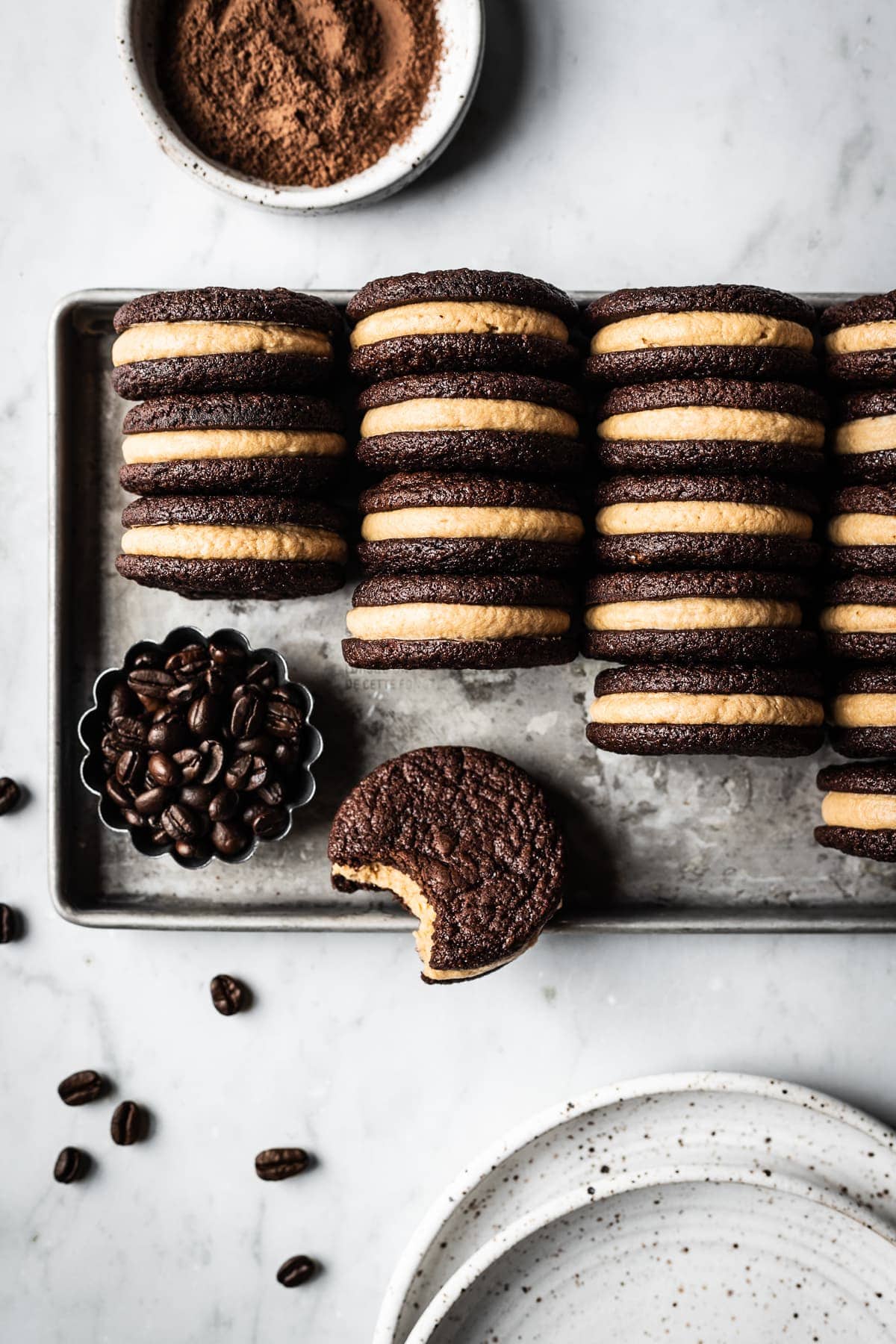 A silver metal cookie sheet holds four tidy rows of frosting filled sandwich cookies on end to show their centers. One is lying flat with a bite taken out of it. The cookie sheet is on a white marble surface. Two speckled white ceramic plates, a small fluted container of coffee beans, and a small white ceramic bowl of cocoa powder are on the border of the image.