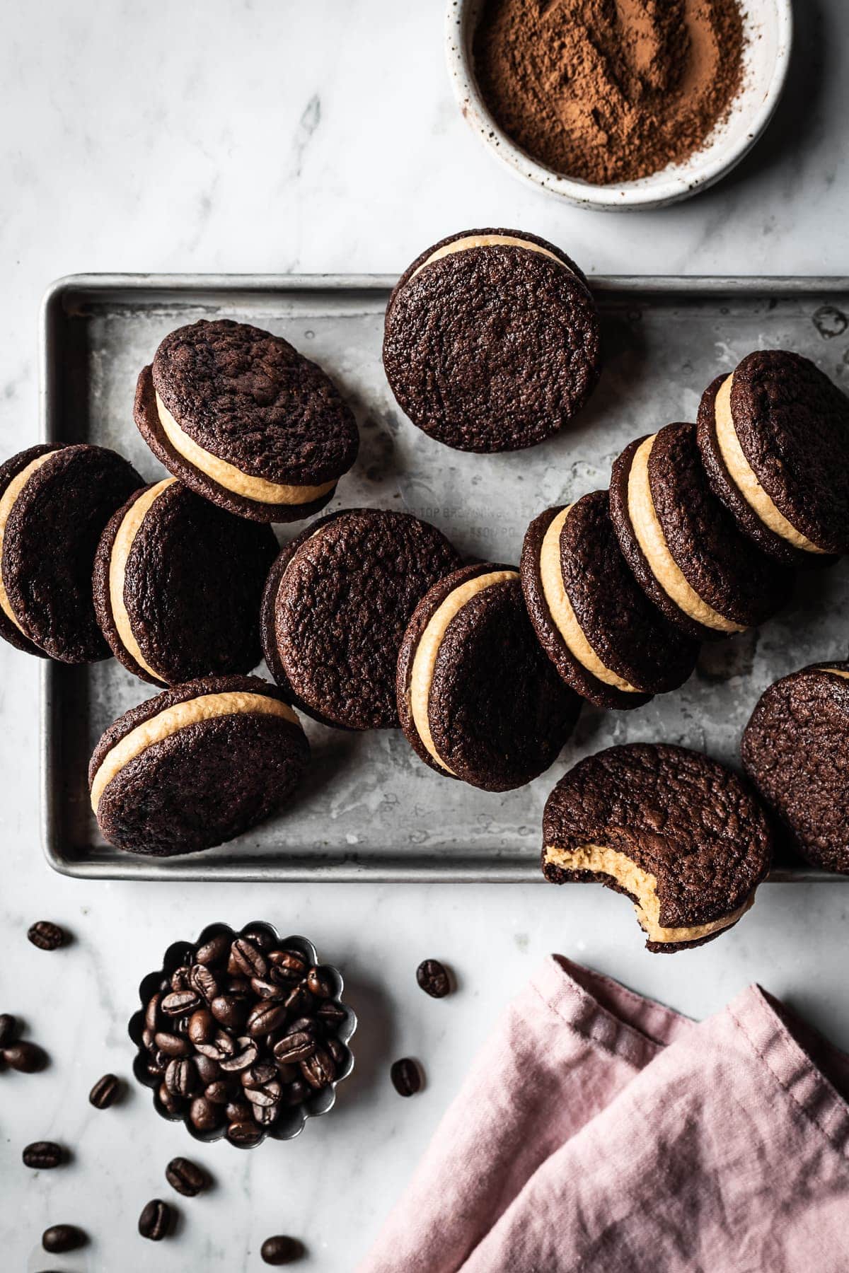 A silver metal cookie sheet holds a messy pile of sandwich cookies, some tipped to reveal their frosted center. The cookie sheet is on a white marble surface. A pink linen napkin, a small fluted container of coffee beans, and a small white ceramic bowl of cocoa powder are on the border of the image.