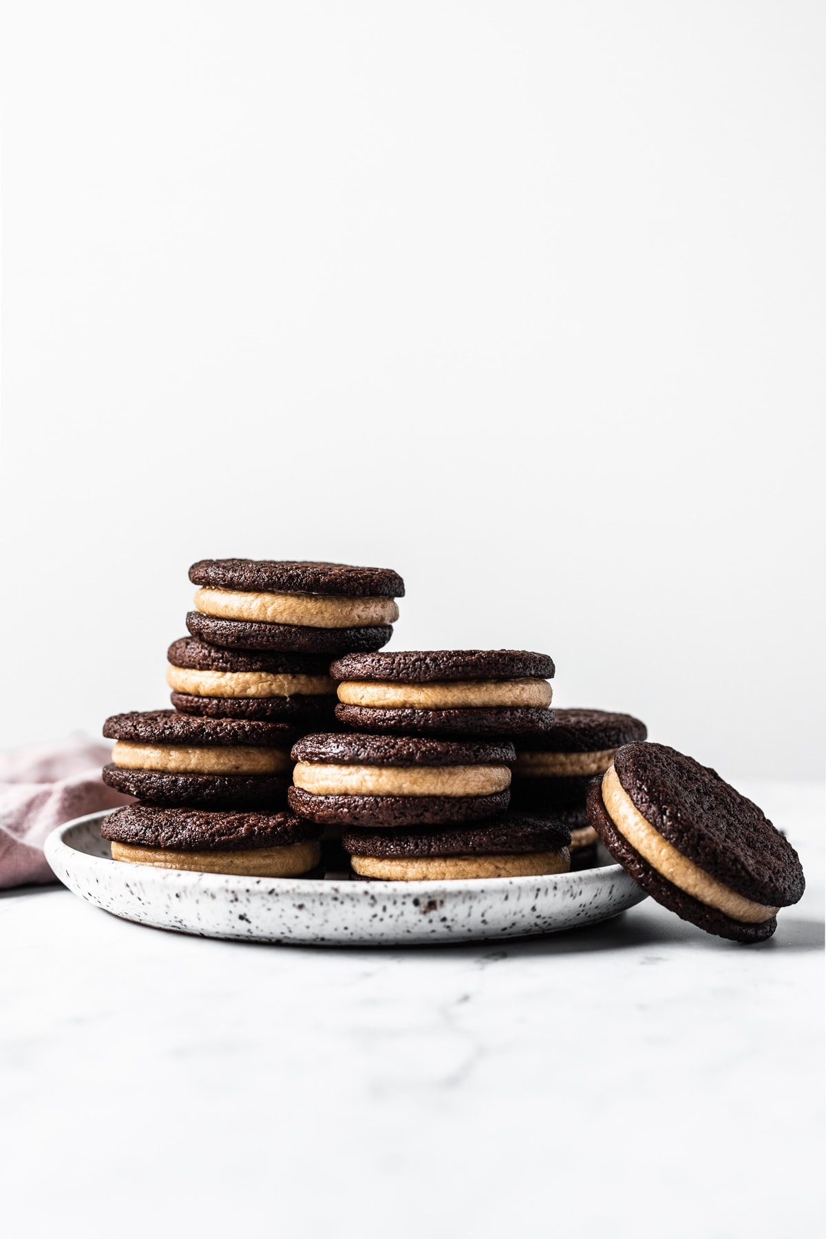 A bright and airy photo of a stack of cookies on a white speckled ceramic plate. A pink linen napkin is folded underneath on a white marble surface. One cookie rests at an angle next to the plate.