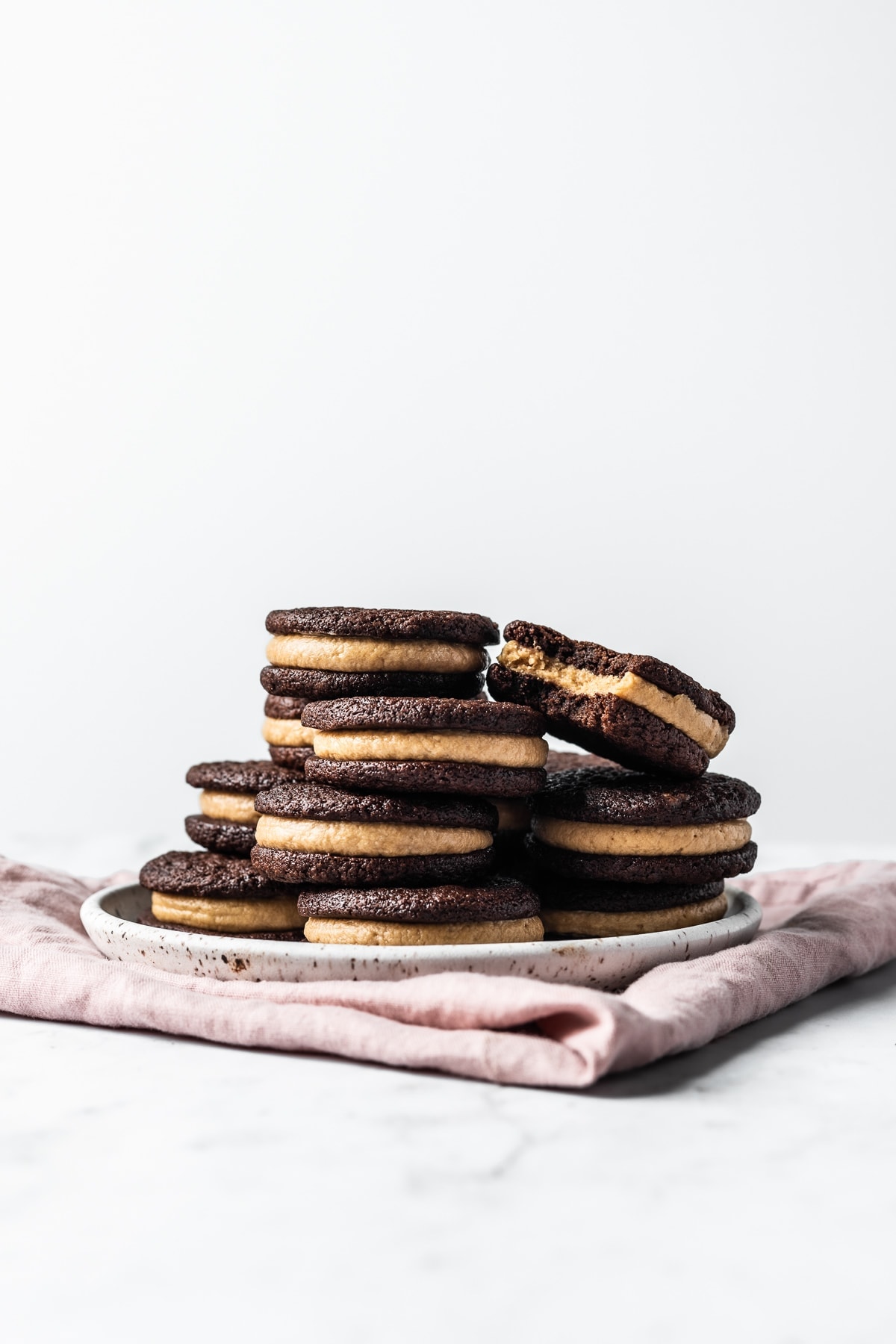 A bright and airy photo of a stack of cookies on a white speckled ceramic plate. A pink linen napkin is folded underneath on a white marble surface.