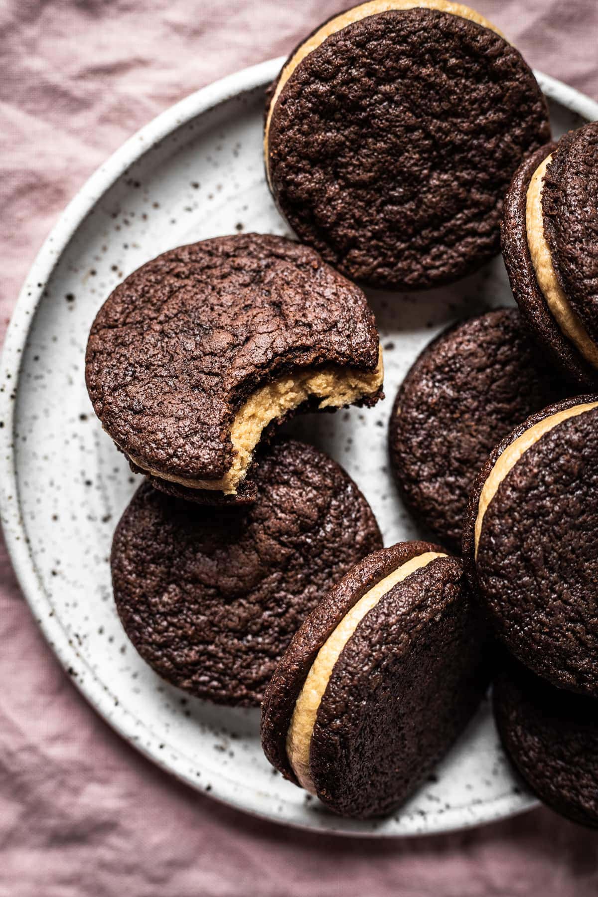 A pile of chocolate cookies on a speckled white ceramic plate on a pink linen surface. One cookie has a bite taken from it.
