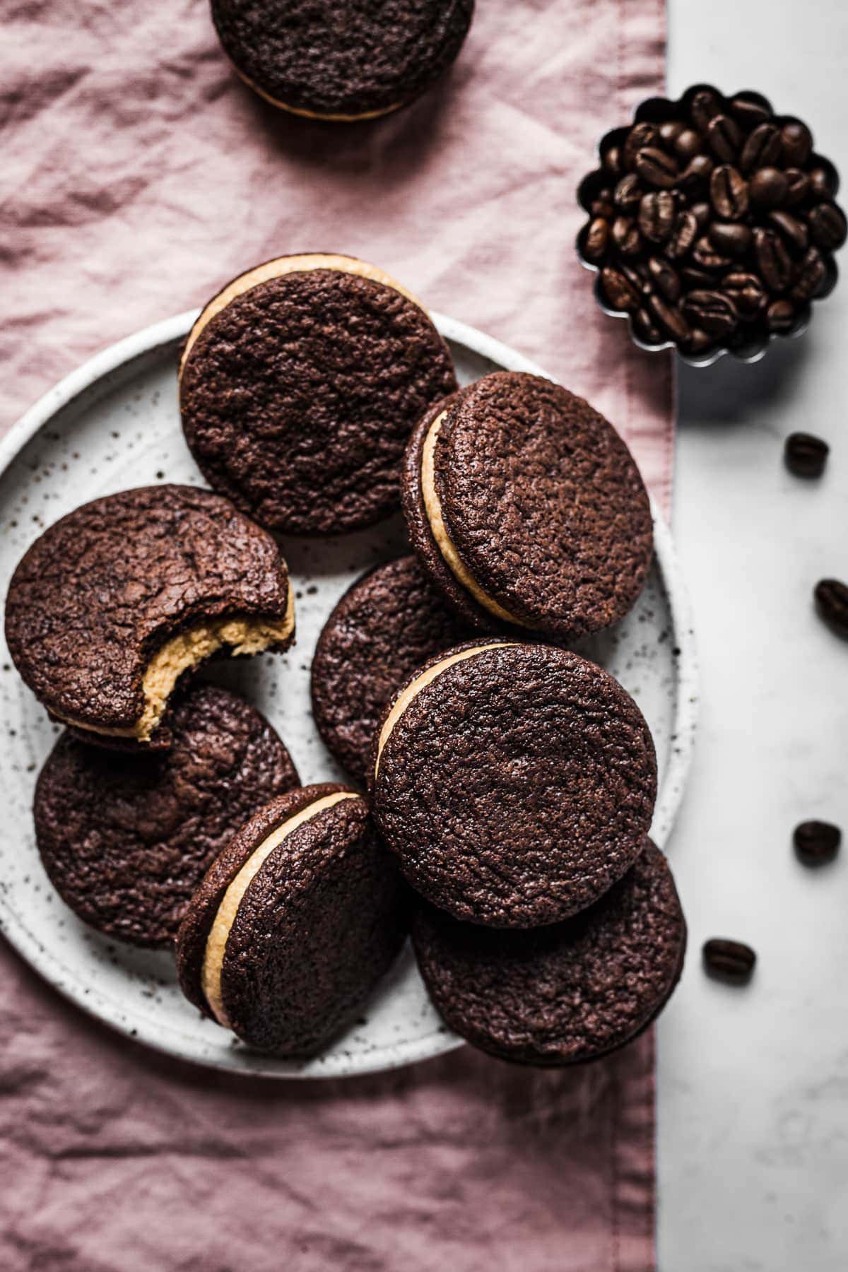 A pile of chocolate cookies on a speckled white ceramic plate on a pink linen surface. Nearby are more cookies and a small fluted container of coffee beans. One cookie has a bite taken out of it, revealing the coffee mascarpone frosting.